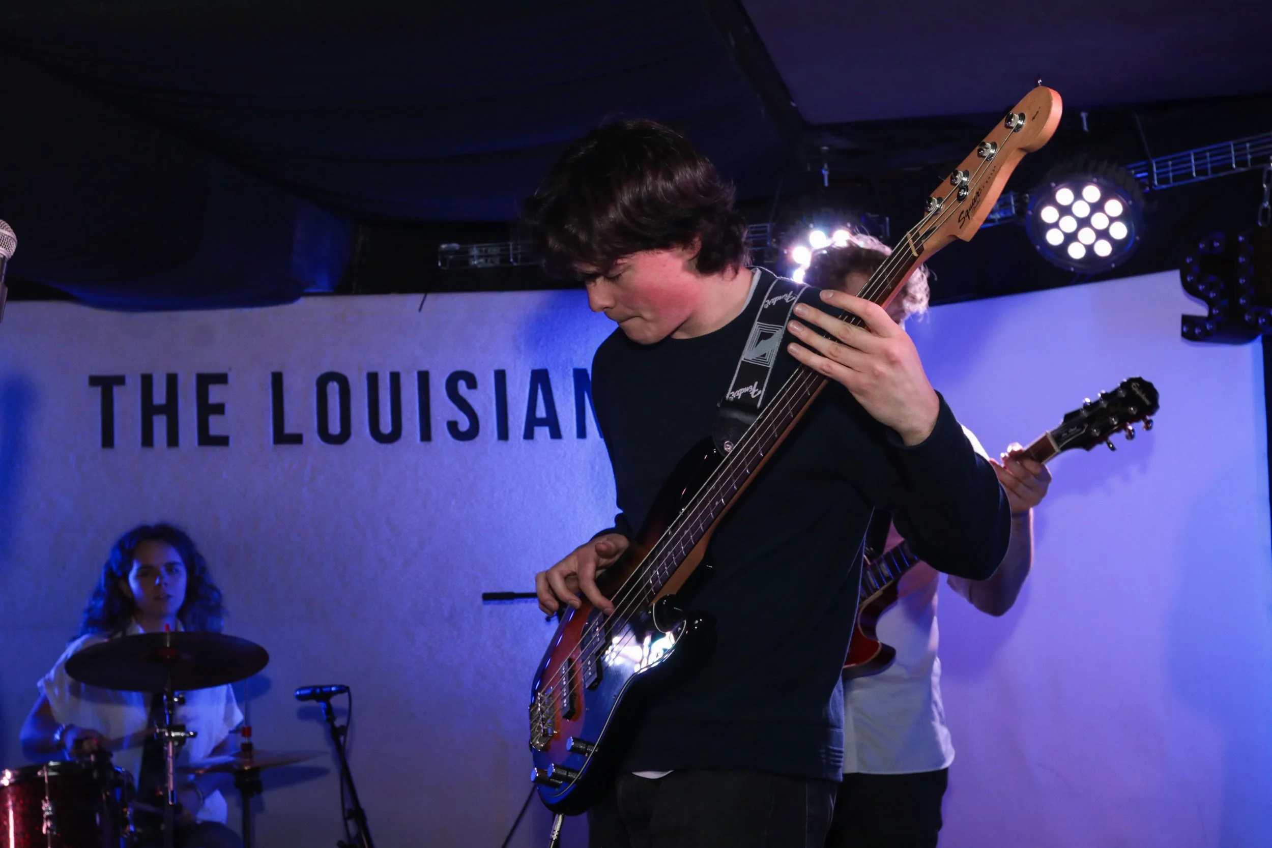 Young man playing the bass guitar during a performance with a woman playing drums in the background at The Louisiana venue.