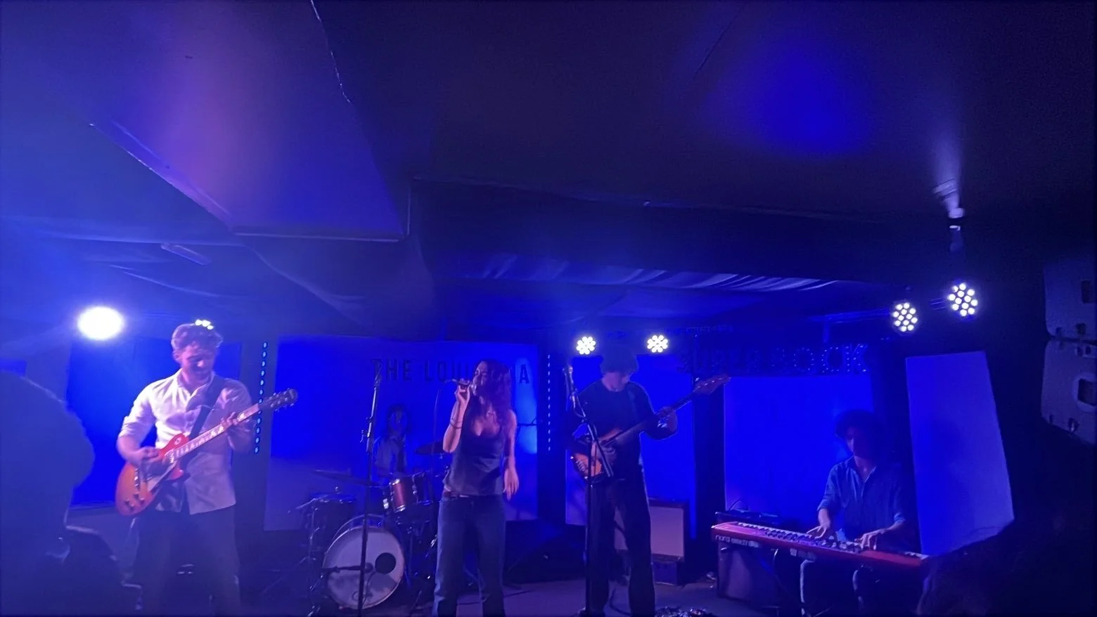 A band performing on stage with blue lighting, featuring a guitarist, singer, bassist, drummer, and keyboardist.