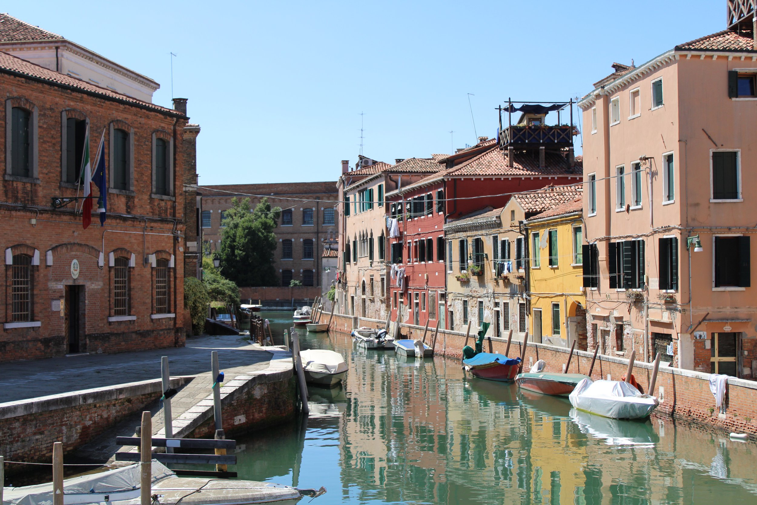 Colorful buildings along a canal with small boats docked, in a sunny Venetian street scene.