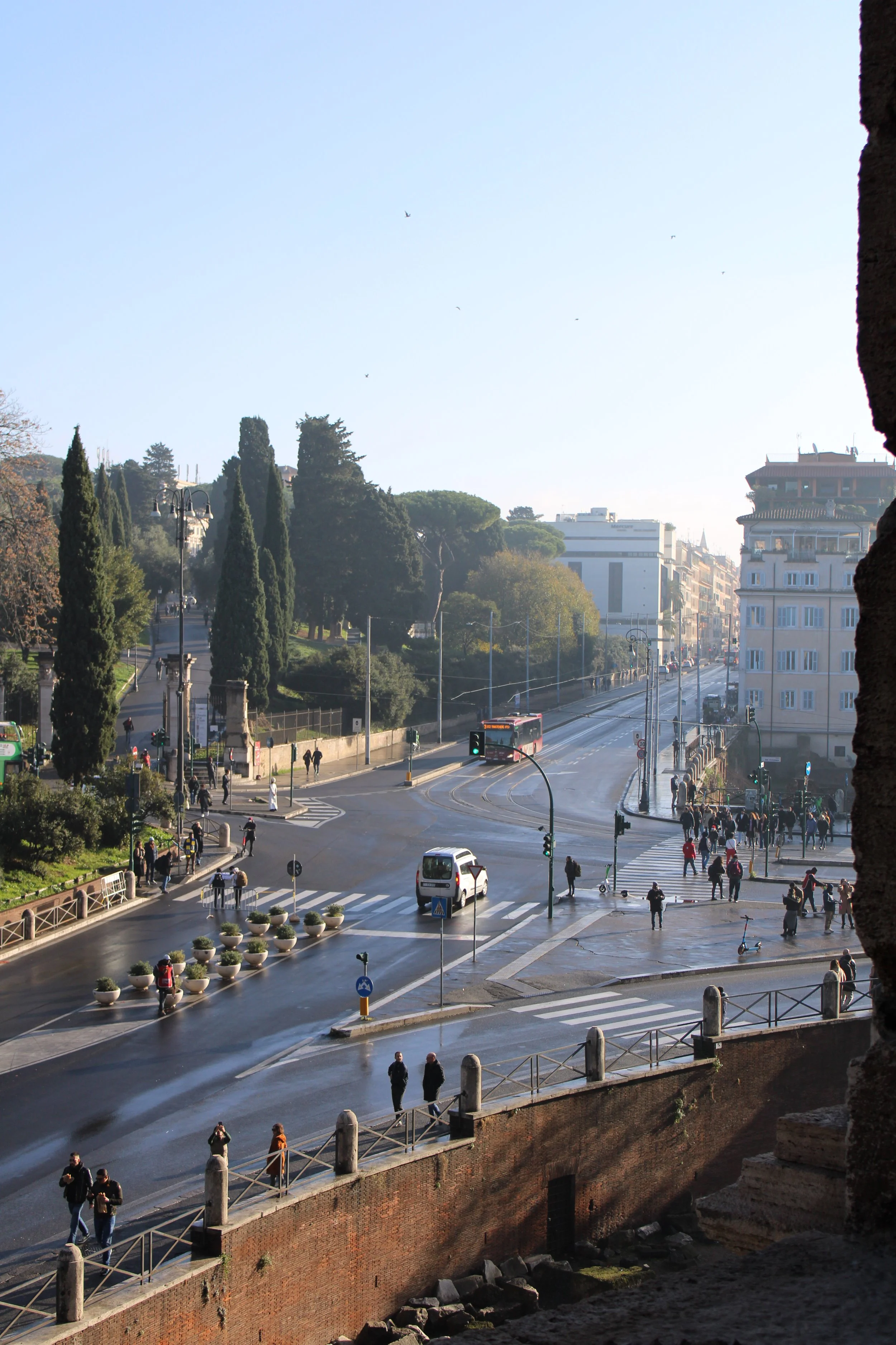 A city street scene showing pedestrians crossing the road, a bus on the street, cars, and trees in the background with buildings and a clear sky.