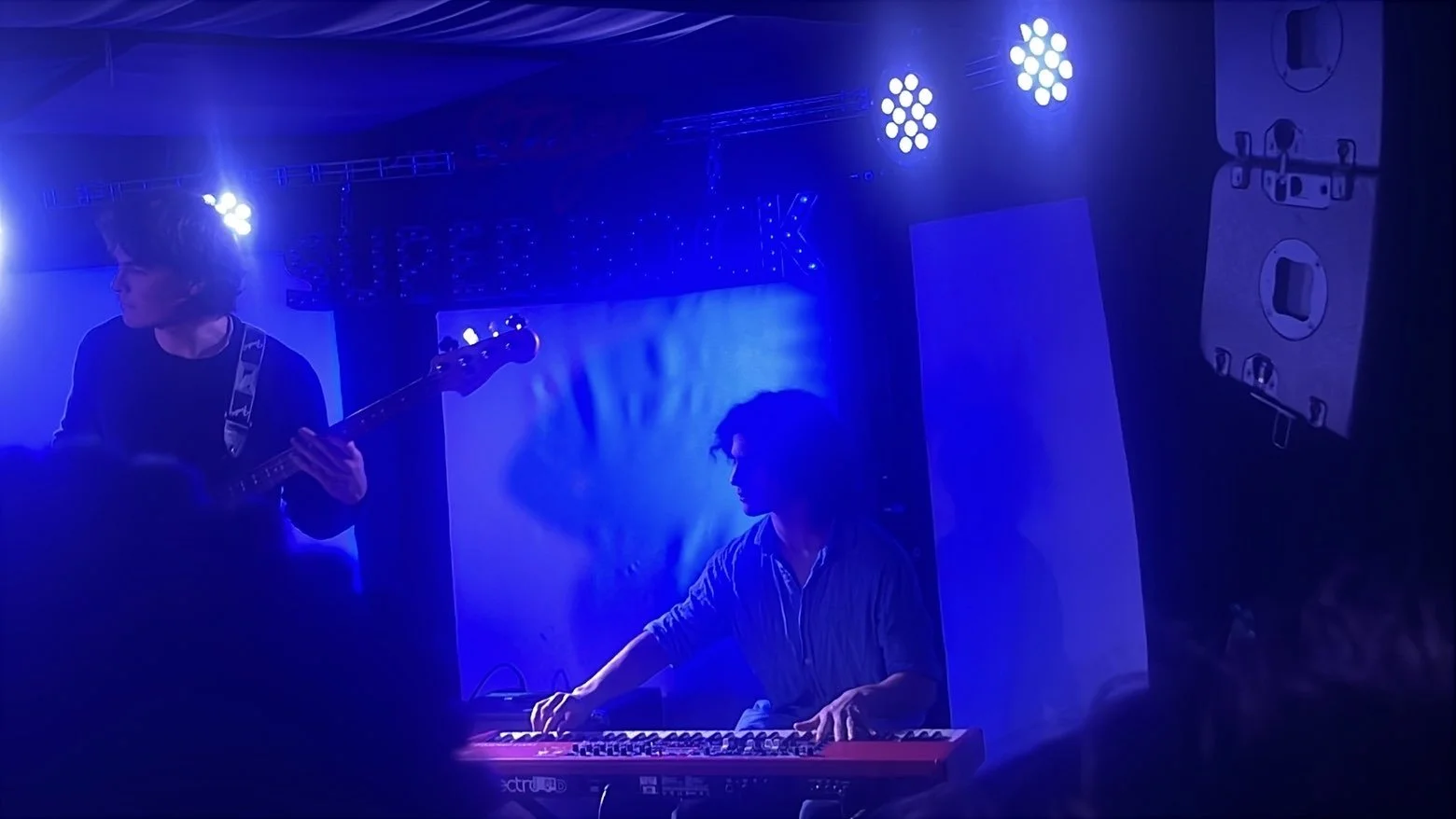Two musicians performing on stage with blue lighting; one playing a bass guitar and the other playing a red keyboard.