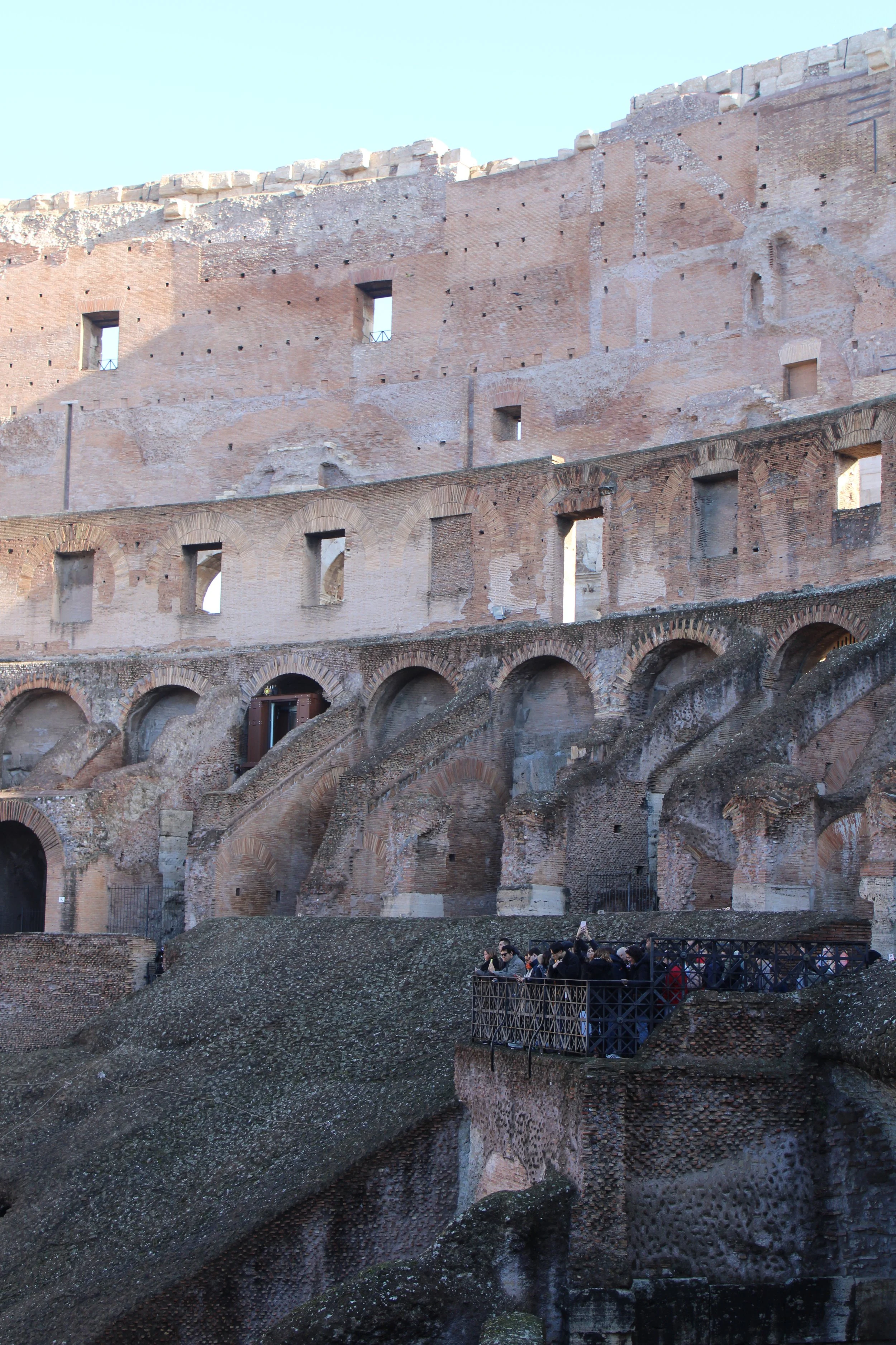 View of the inner walls of the ancient Roman Colosseum showing multiple levels of arches and small windows, with a group of tourists on a viewing platform at the bottom right.