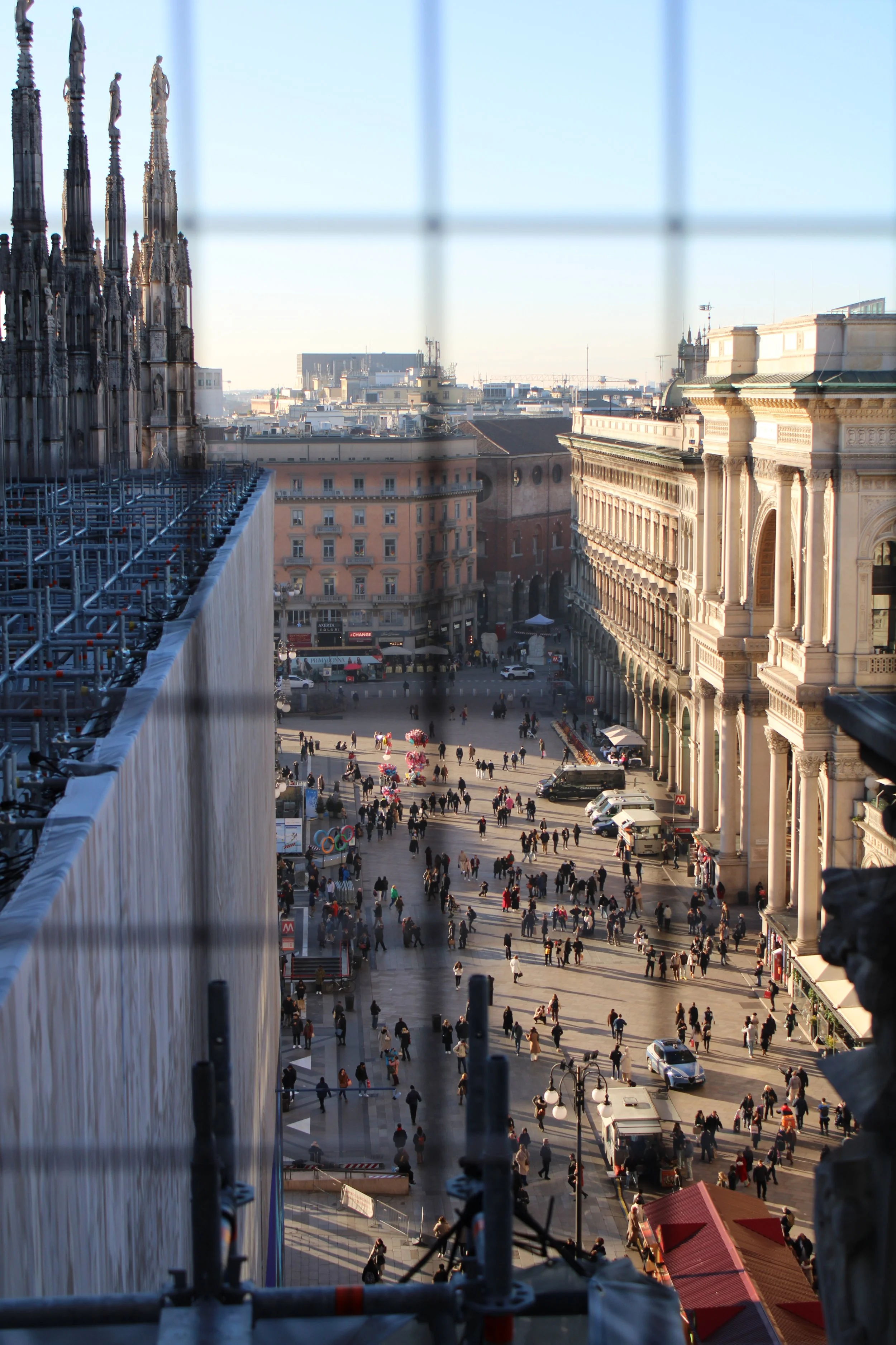 Aerial view of a busy city square surrounded by historic buildings with numerous pedestrians and vehicles, framed through a wire mesh.
