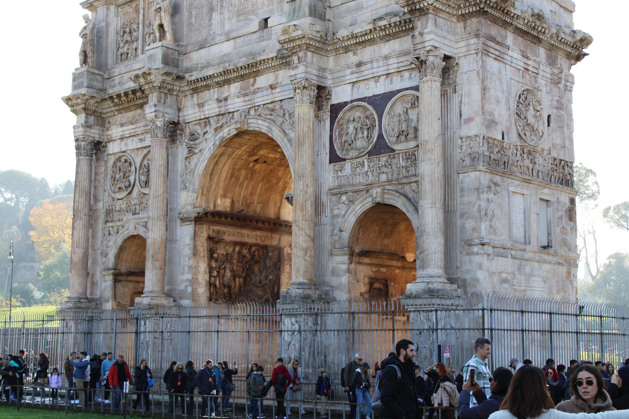 People standing and walking near the Arch of Constantine in Rome, Italy, with trees and a clear sky in the background.
