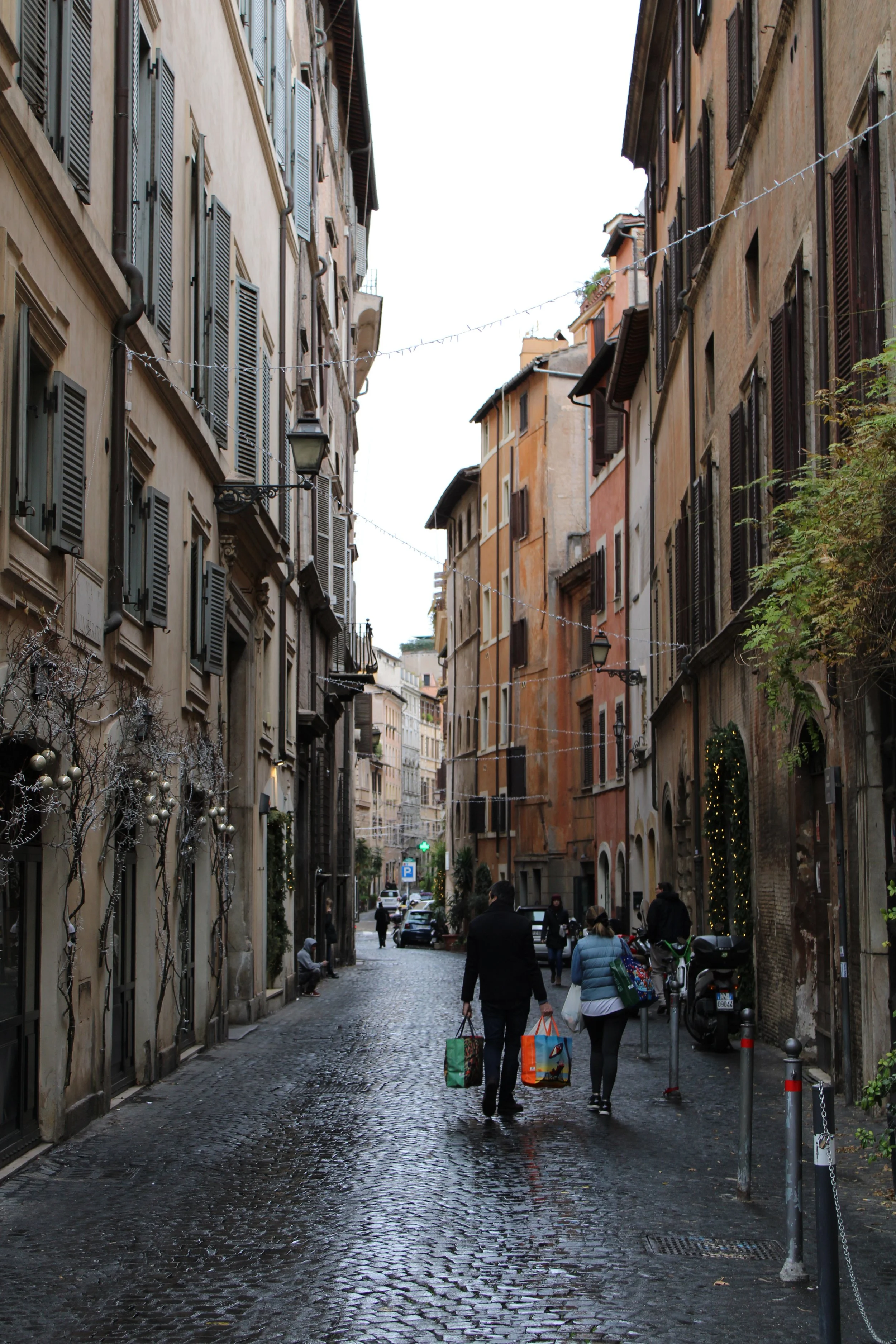 A narrow cobblestone street in a European city with tall historic buildings on both sides decorated with string lights and holiday ornaments, with people walking and carrying shopping bags.
