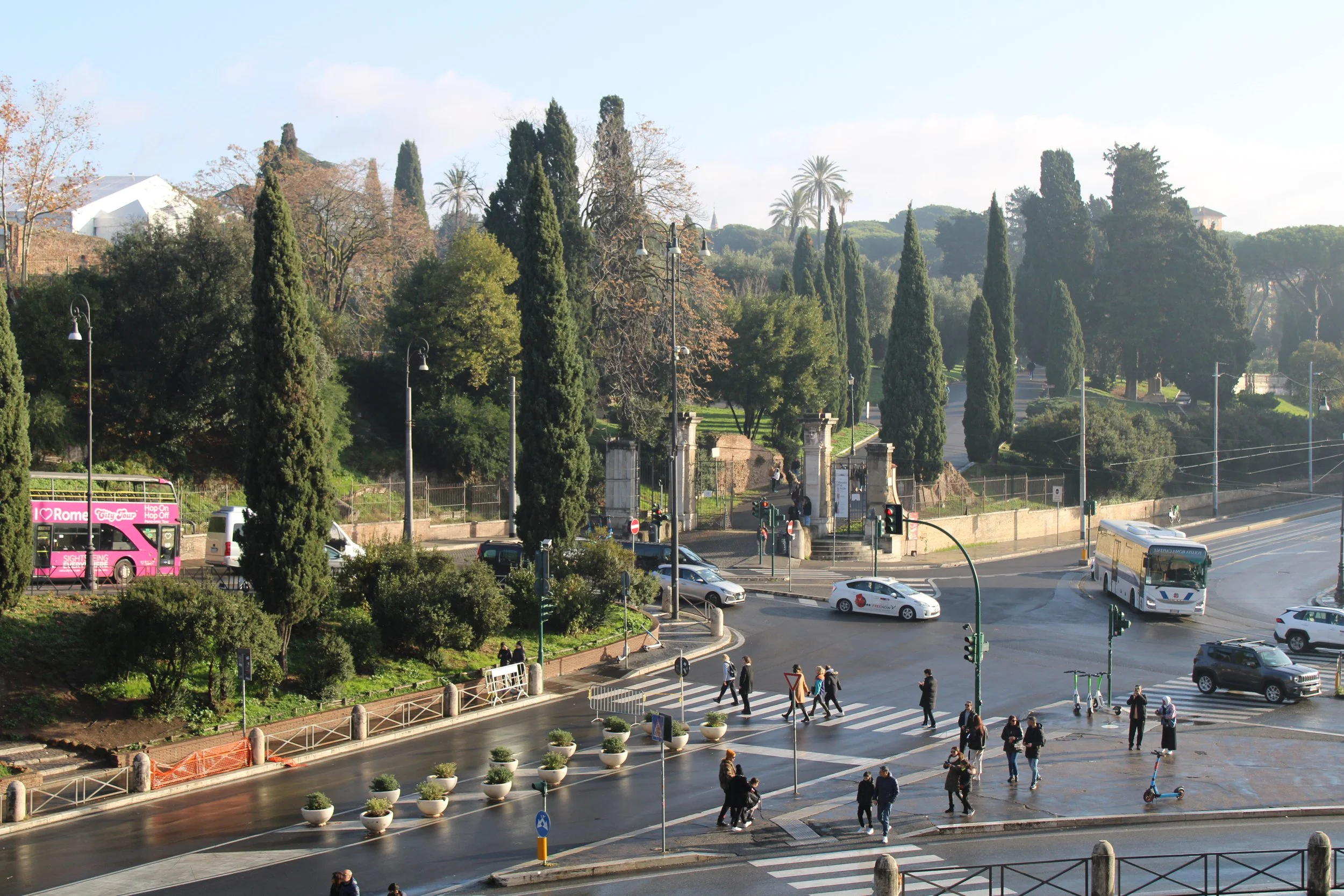 Street view of a busy intersection with pedestrians crossing, cars and buses, and a park with tall trees in the background.