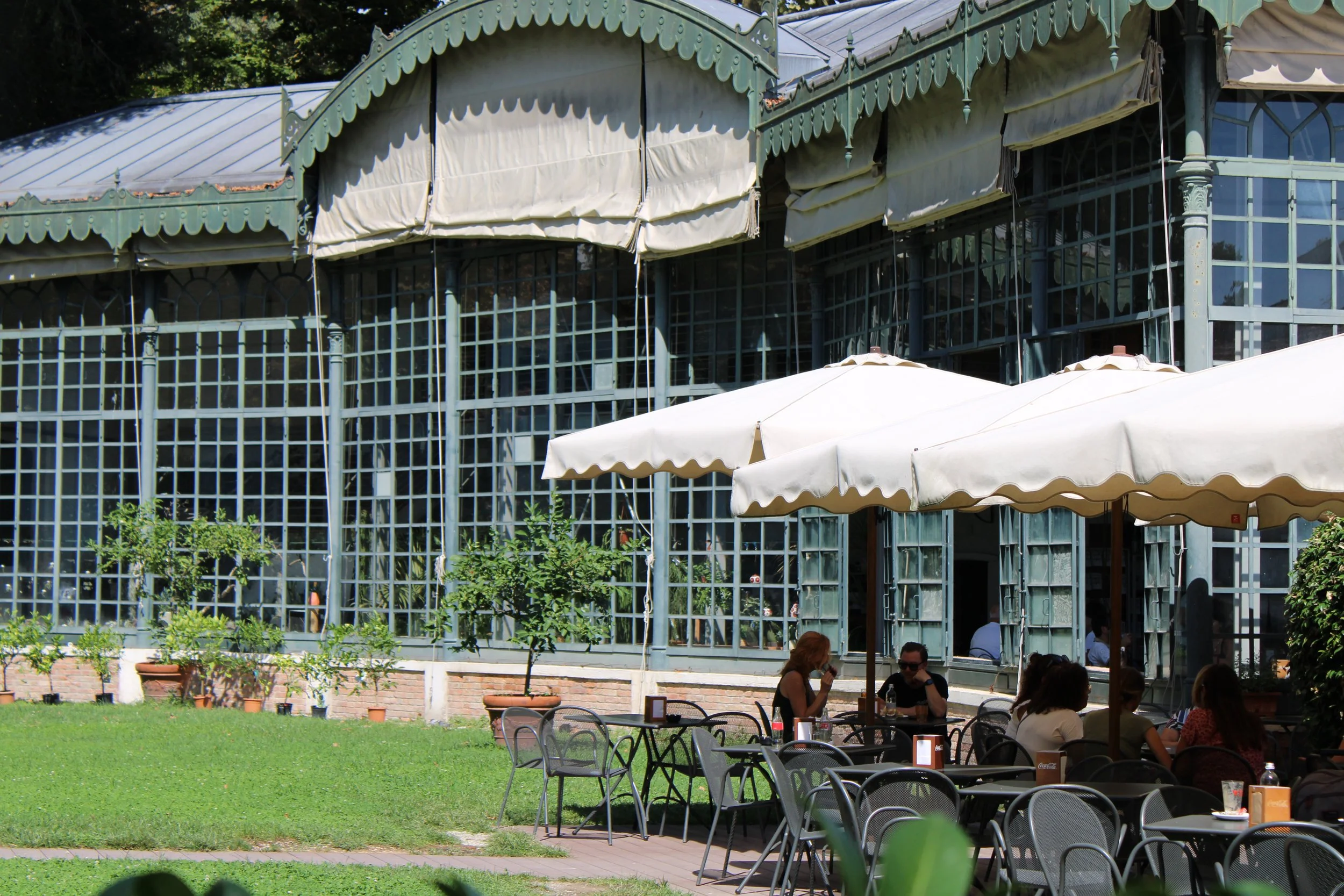 Outdoor dining area with tables, chairs, and large umbrellas, in front of a glass greenhouse structure, with people sitting and dining.