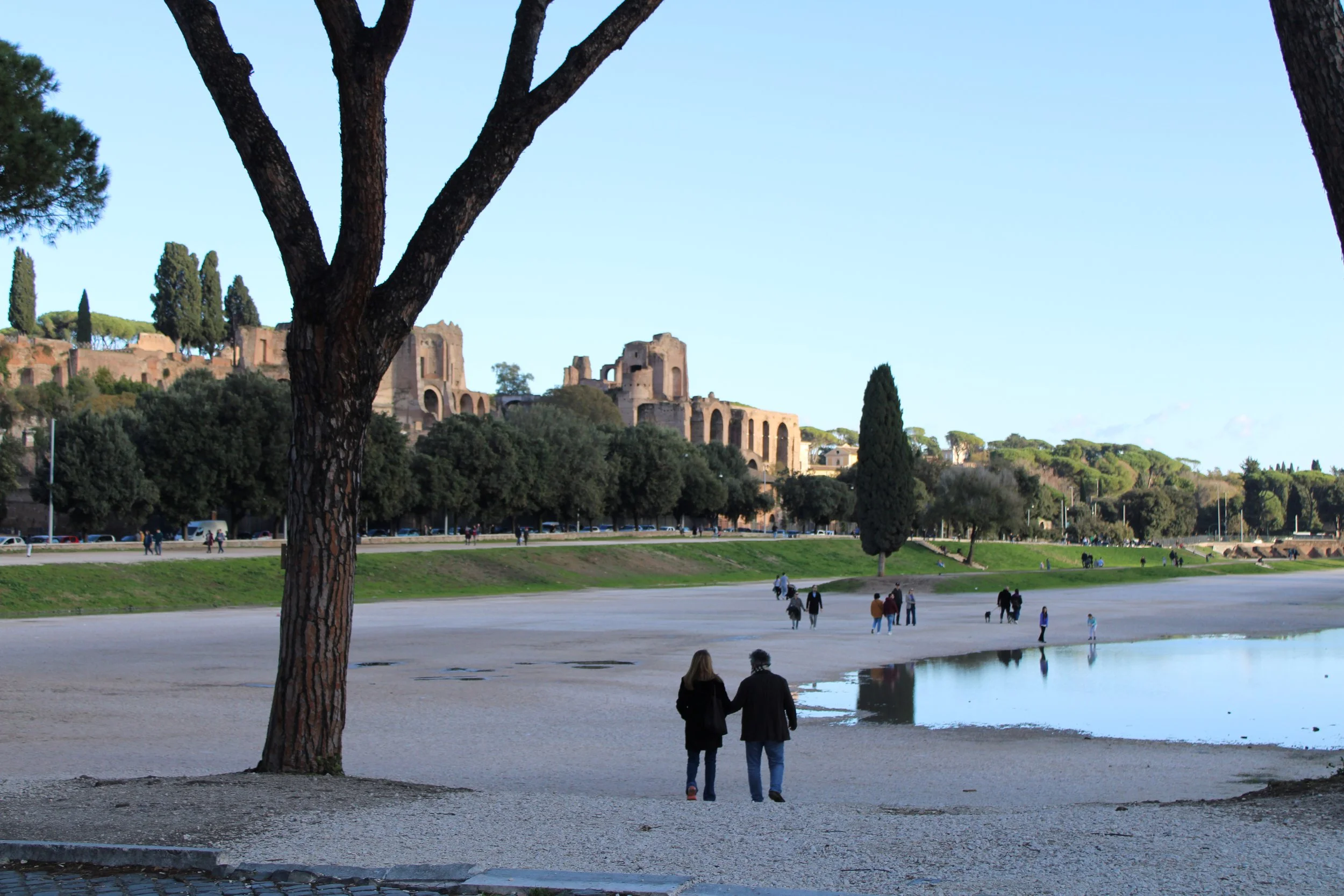 People walking along a sandy beach with a small pond, trees, and ancient ruins in the background under a clear sky.