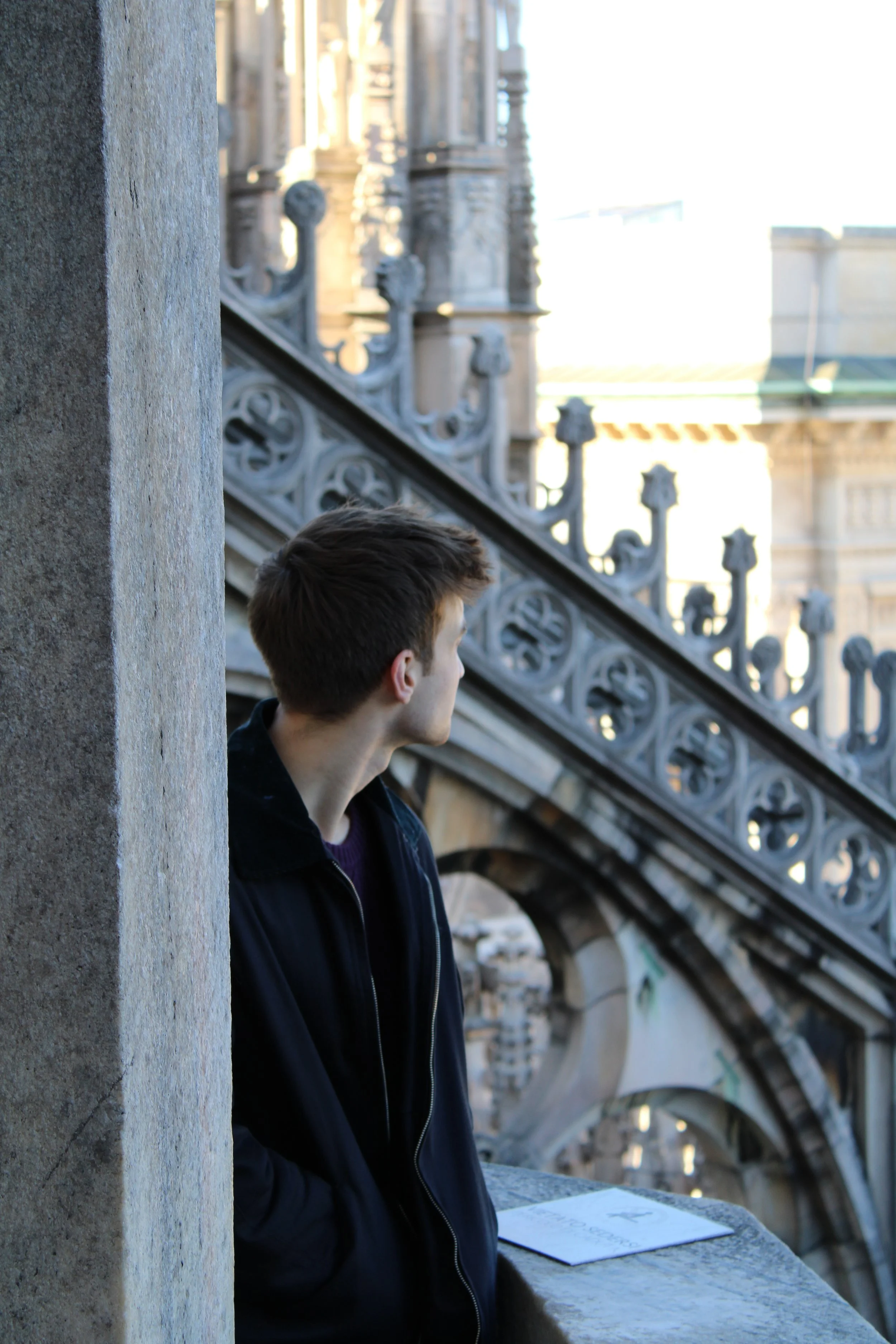 A young man with brown hair wearing a black jacket looks out over a cityscape from a historic building's balcony, with ornate architecture visible in the background.