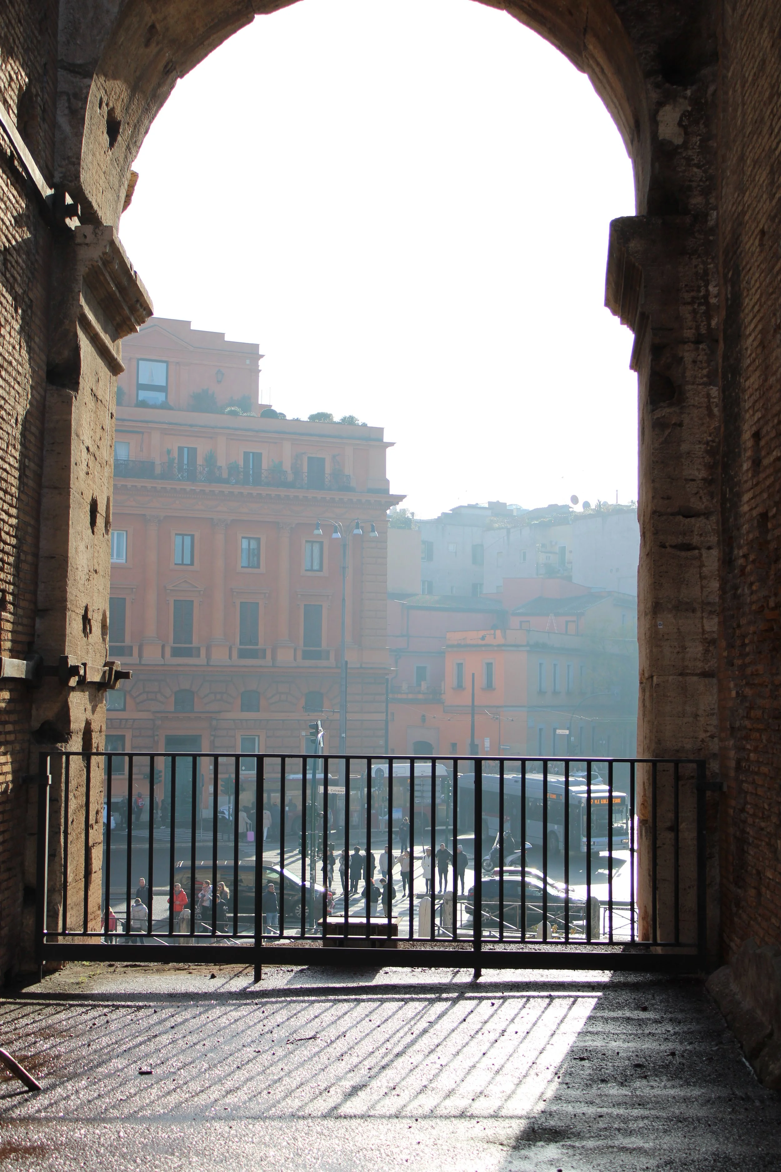 Cityscape viewed through an arched stone window with cast shadows on the ground, showing buildings, cars, and pedestrians on the street.