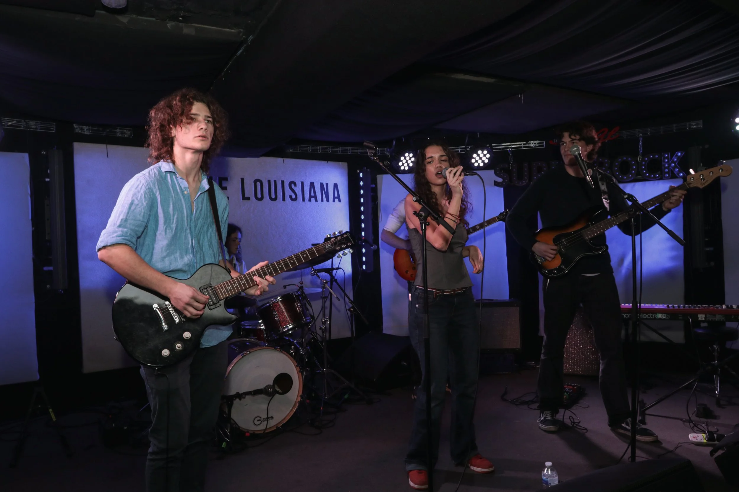 A band of four young musicians performing on a stage in Louisiana. The band includes a guitarist, a vocalist, a bassist, and a drummer in the background. The stage has a dark ceiling and illuminated background with signs.