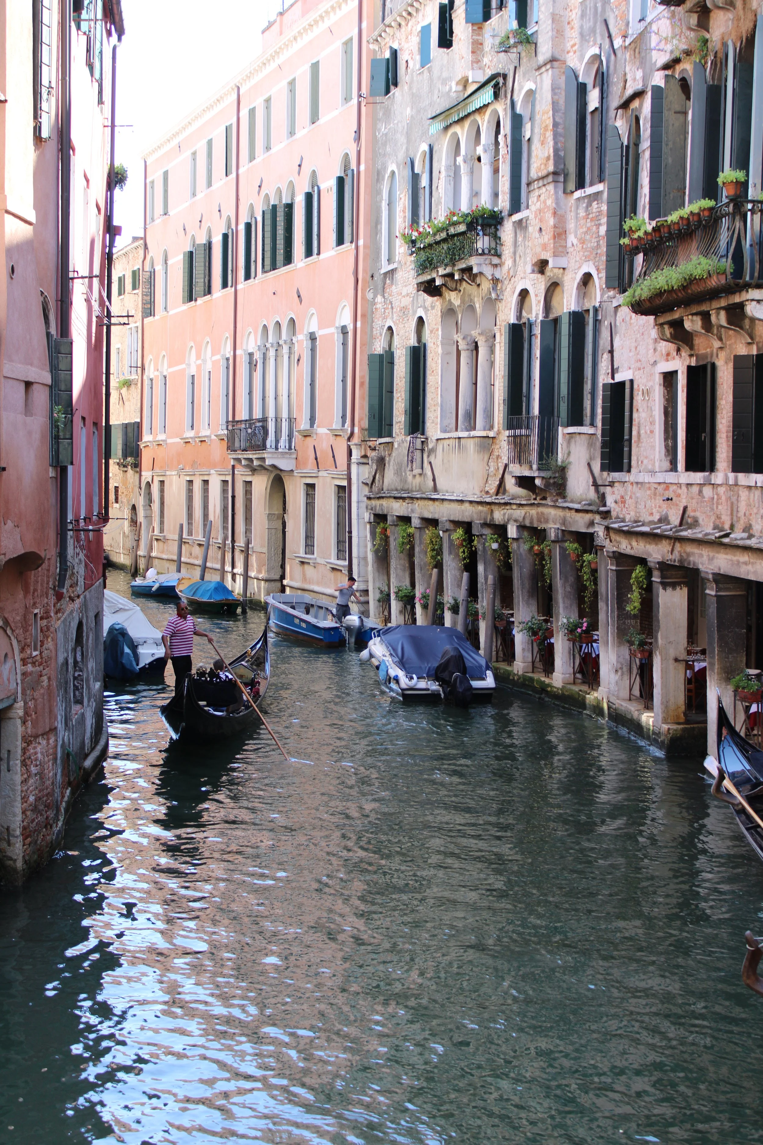 A canal in Venice with several boats, including a gondola, and tall historic buildings with shuttered windows and balconies.