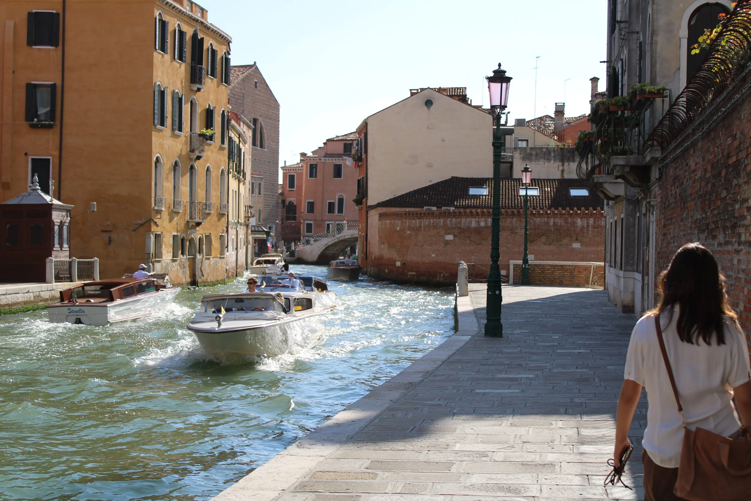A canal in Venice, Italy, with boats sailing past colorful historic buildings, a pedestrian walkway, and a woman walking along the promenade.