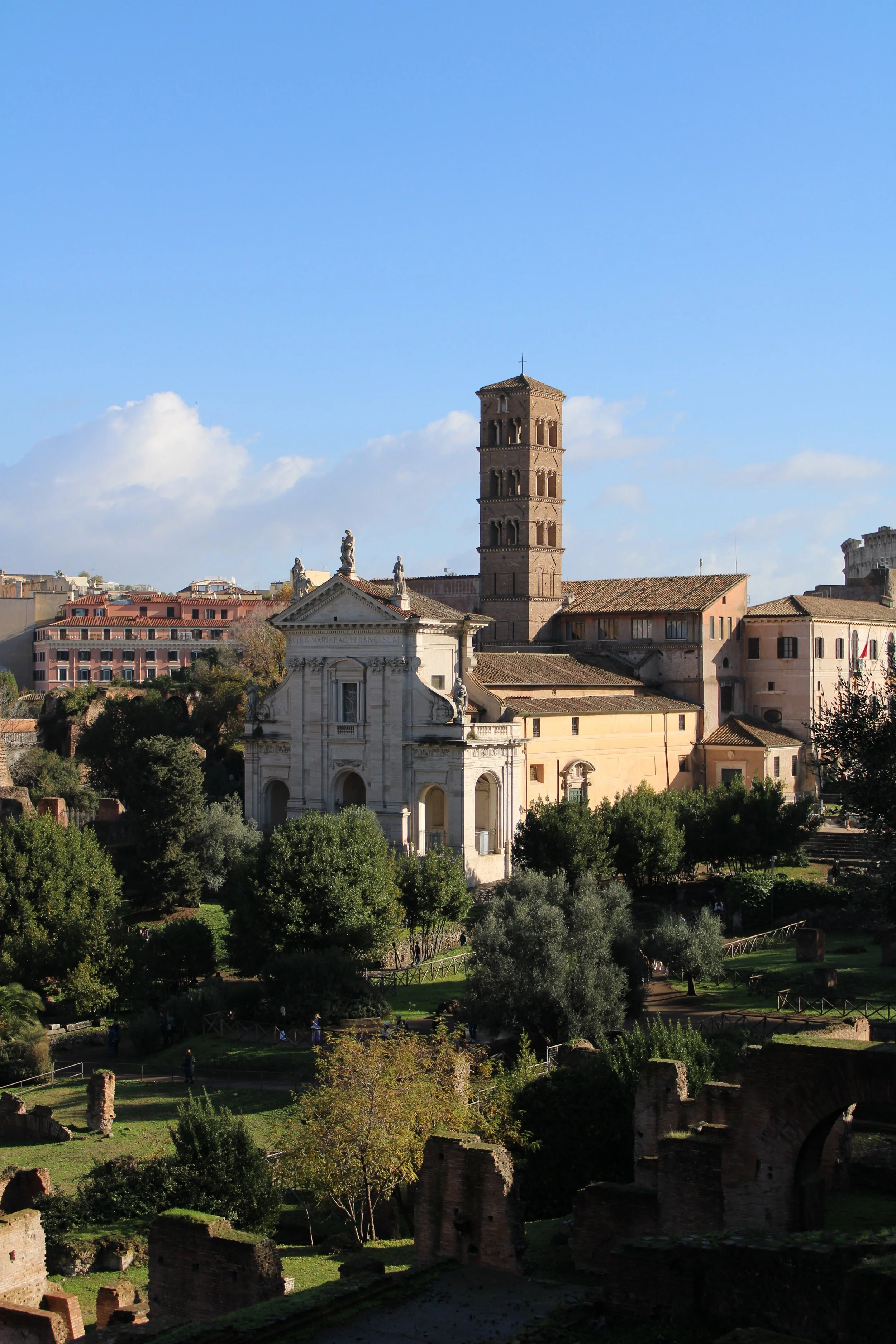 View of ancient Roman ruins with trees in the foreground and historic buildings including a tall brick tower in the background, under a partly cloudy sky.