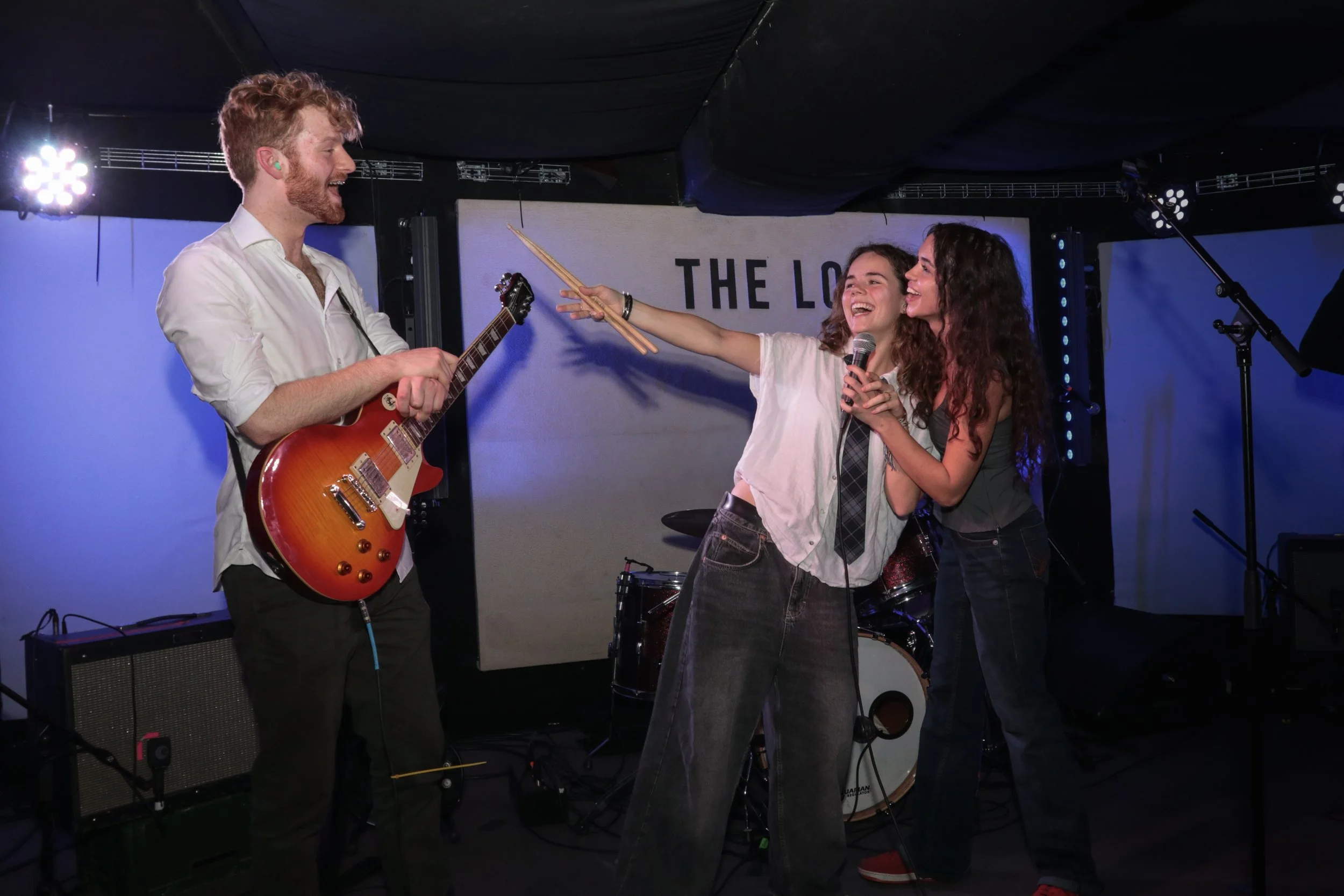 Three young musicians performing on stage, two women and one man. The man is playing an electric guitar and smiling, while the women are holding a microphone and a pair of drumsticks, smiling and interacting cheerfully.
