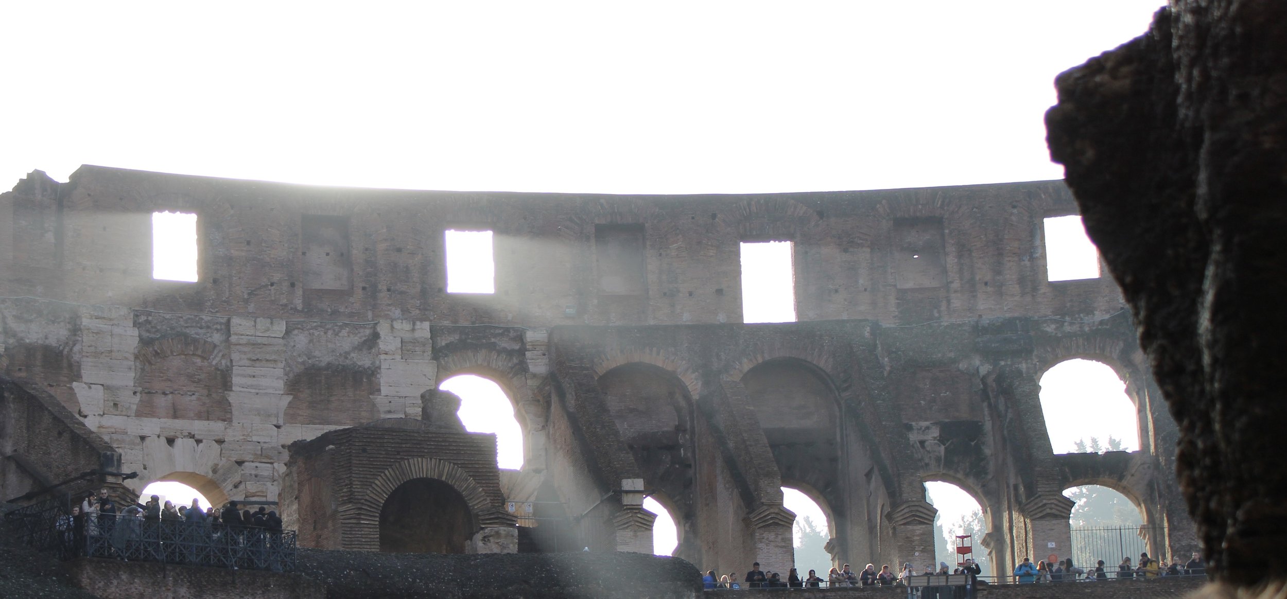 The interior of the Colosseum in Rome with sunlight streaming through large openings and visitors sitting and walking around.