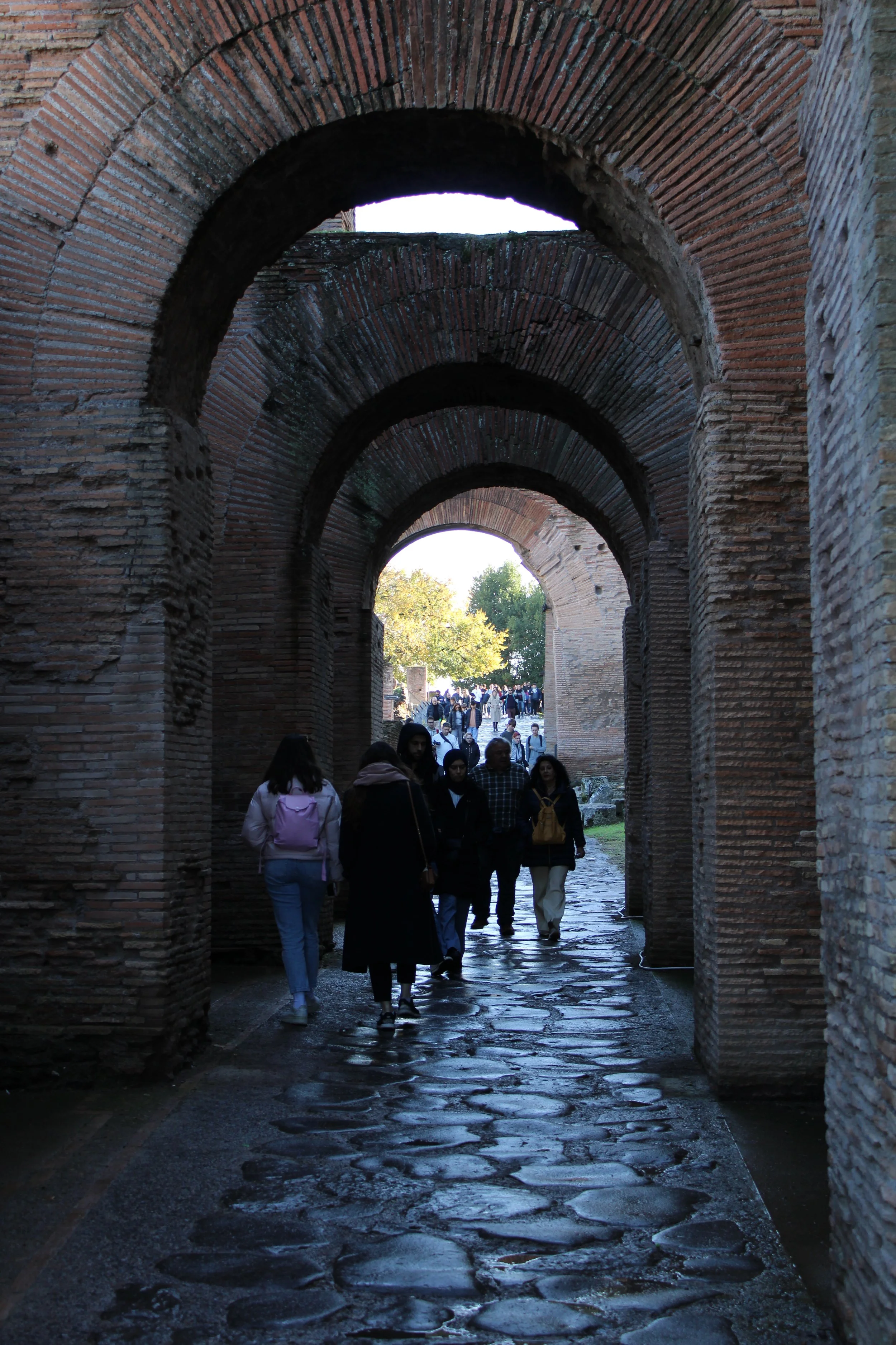People walking through ancient brick archways with rain-soaked cobblestone pathway.
