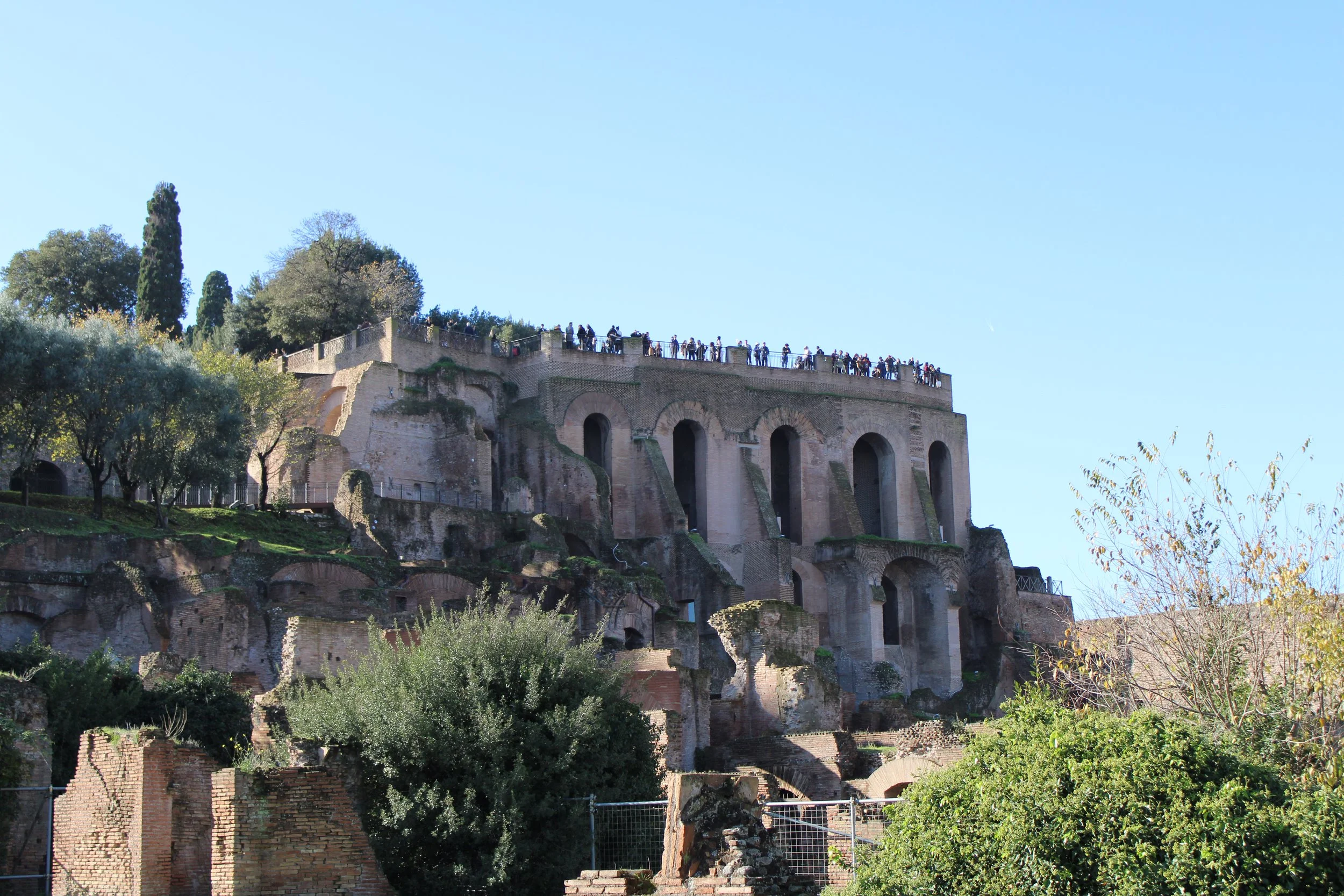 Ancient ruins of a large, multi-level stone structure on a hilltop, with arches and stairs, surrounded by trees, under a clear blue sky