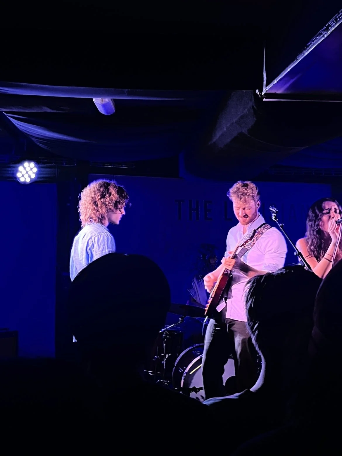 Musicians performing on a dimly lit stage, with one person playing a guitar and others singing, in front of an audience.