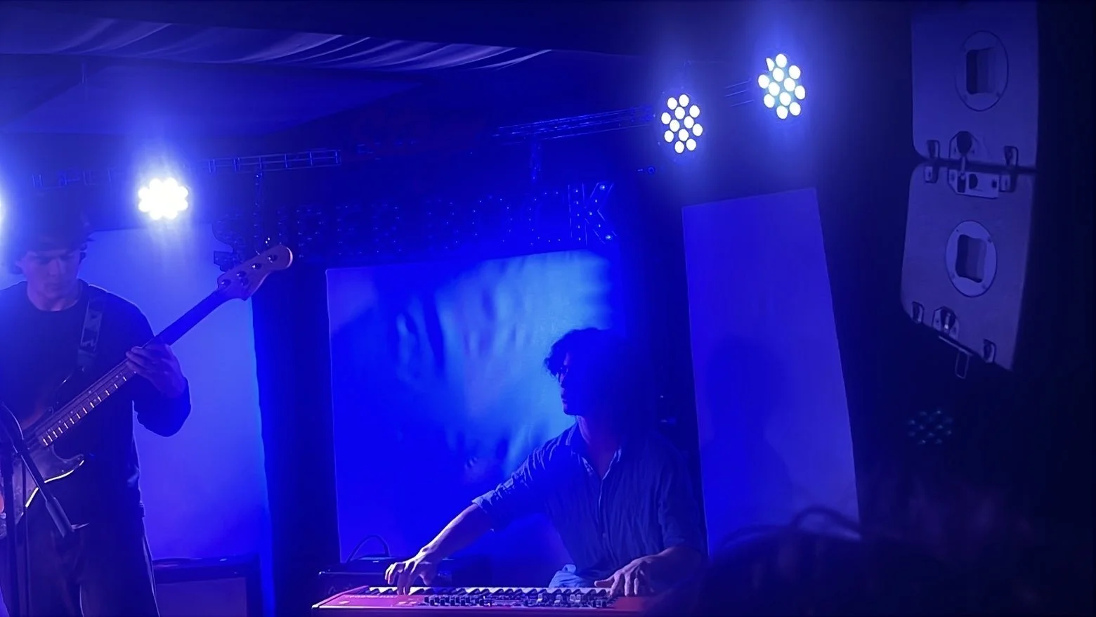 Two musicians performing on stage with blue lighting, one playing bass guitar and the other using a keyboard.