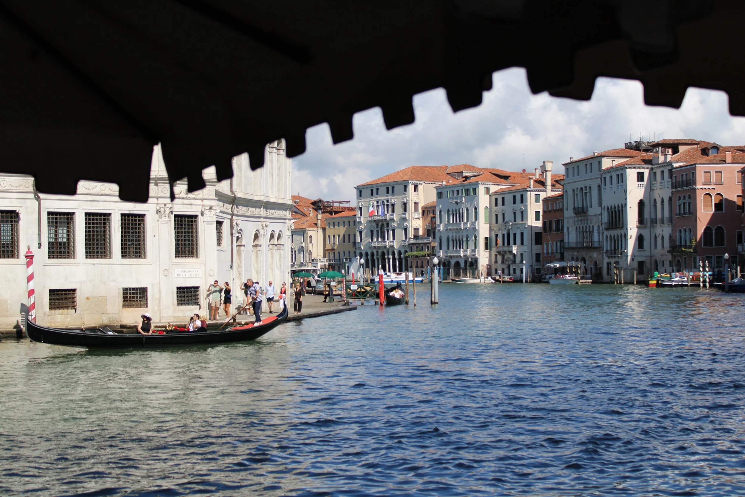 View of a canal in Venice, Italy with colorful buildings along the water, people walking by the water, gondolas, and boats, seen from under a bridge canopy.