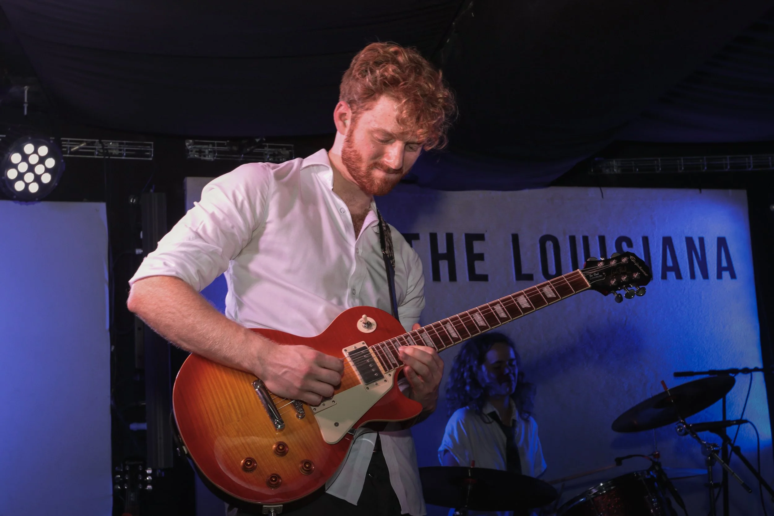 A man with curly red hair and a beard playing an orange electric guitar on stage. There is a woman with curly hair in the background playing drums. The stage has a sign that reads 'THE LOUSIANA'.