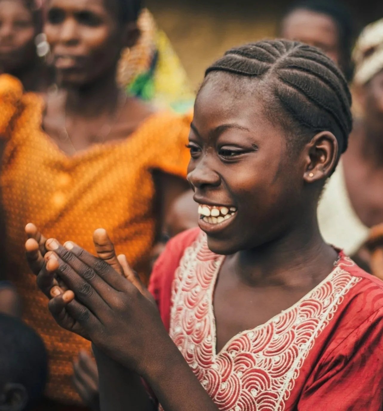 A young African girl is smiling and clapping her hands while other people look on, during a gathering or celebration.