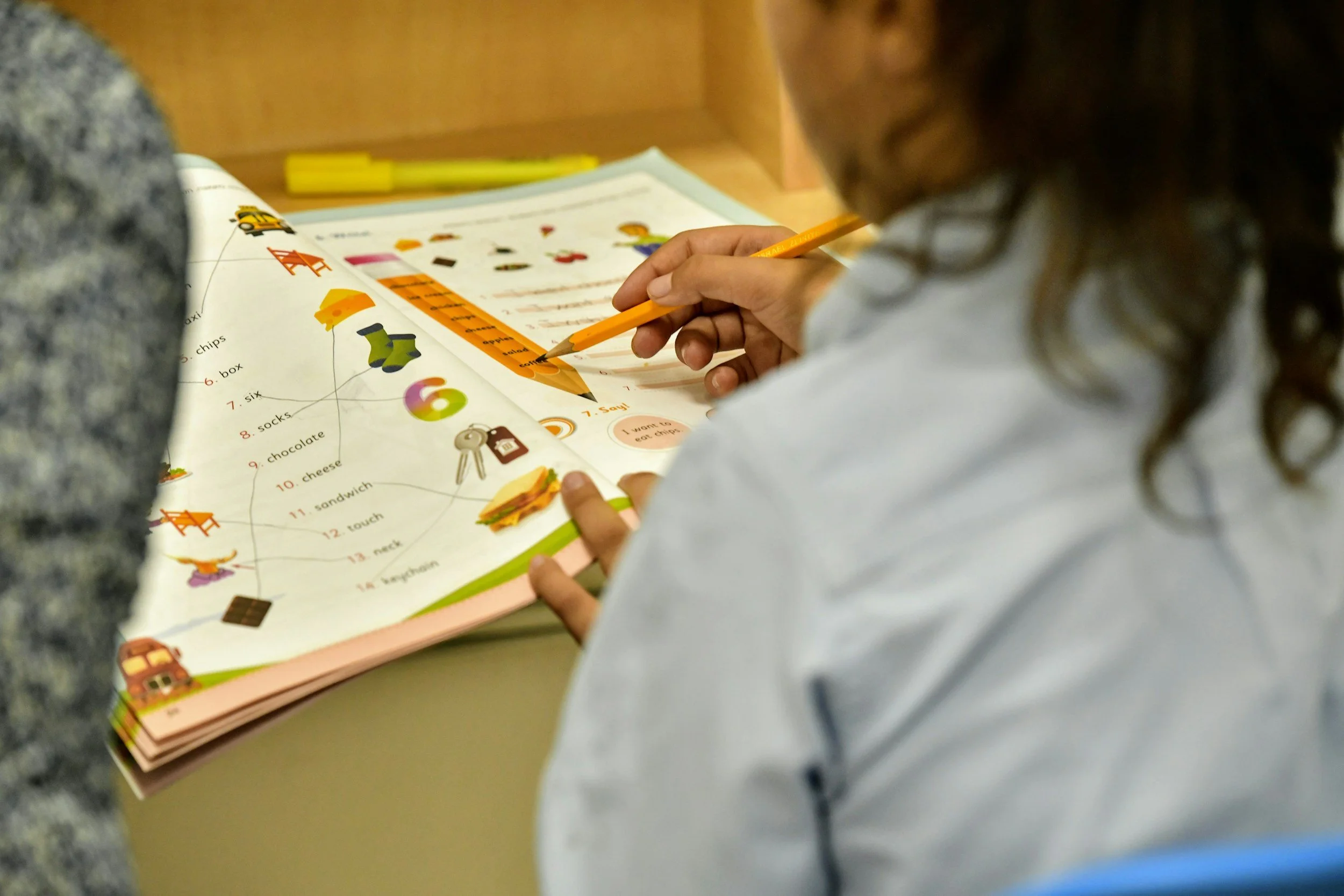 A child is working on a colorful educational workbook that features illustrations of objects, numbers, and words; the child is holding a pencil and pointing to the workbook, which is open on a desk in a classroom setting.