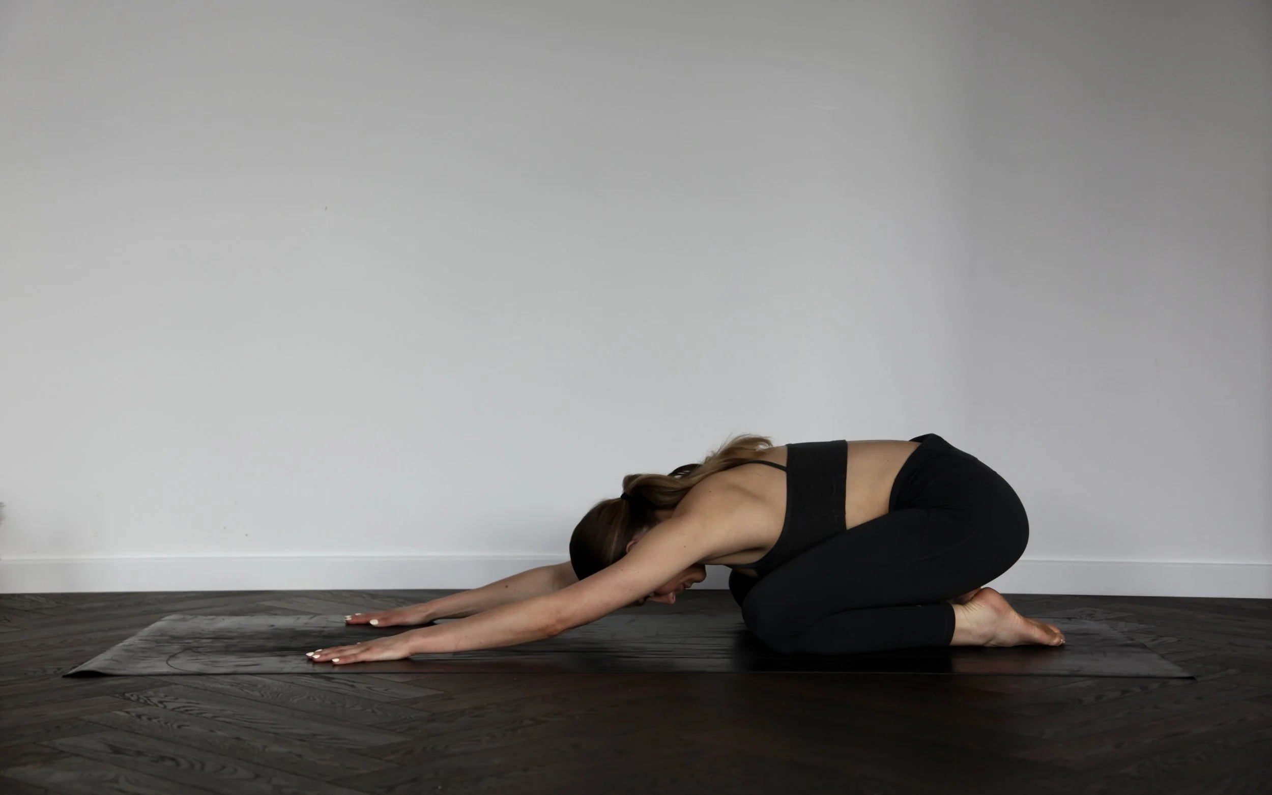 Evie, co-founder of Feast & Flow, practising child's pose on a yoga mat with her eyes closed and a soft smile. She is wearing a black sports bra and matching black leggings in a calm, minimal wellness studio setting.