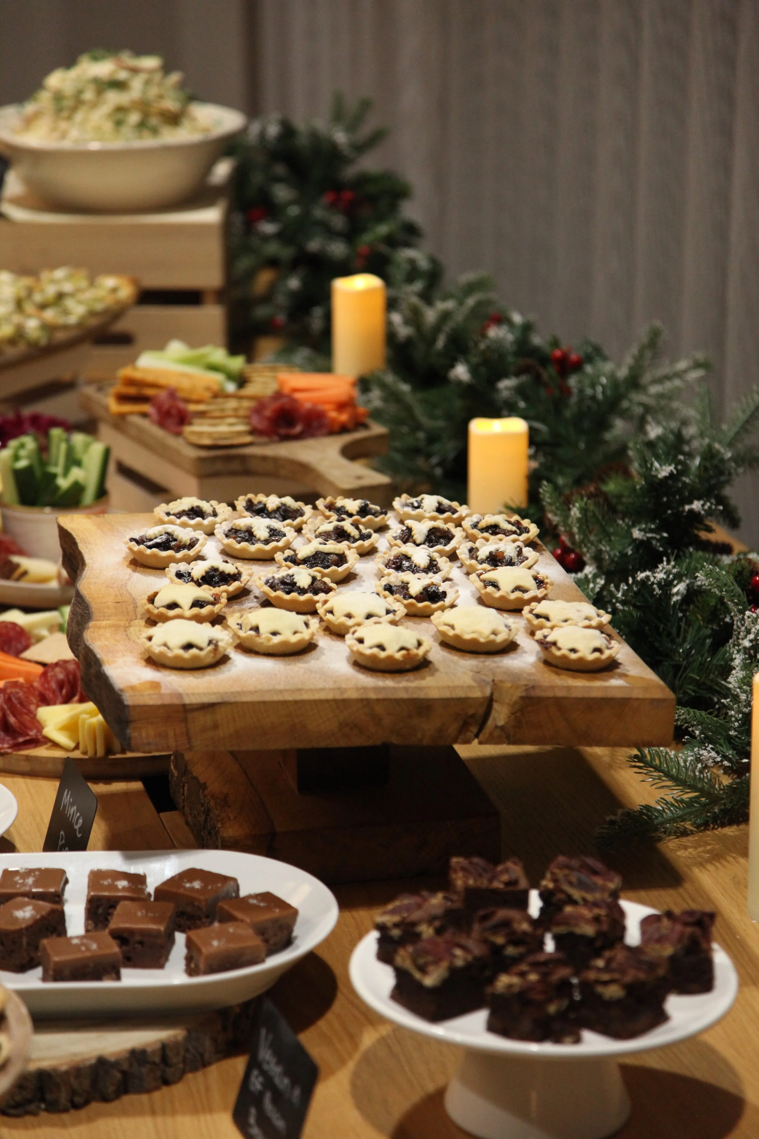 Assorted Christmas desserts and snacks on rustic wooden and ceramic serving dishes, decorated with holiday greenery and candles.