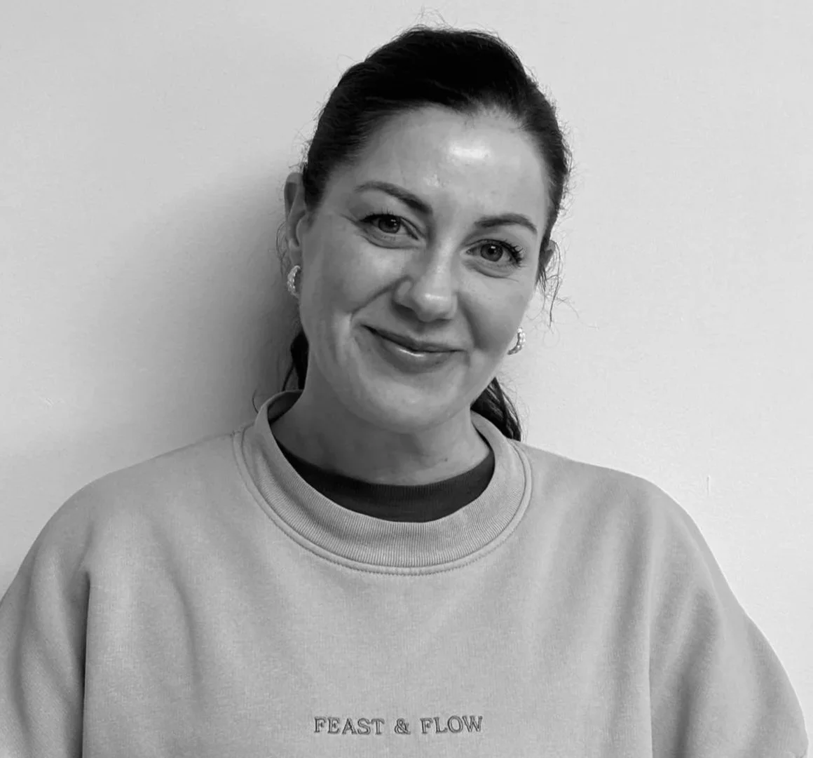 Black and white photo of Mel with dark hair pulled back, smiling and wearing a sweatshirt with 'FEAST & FLOW' written on it, standing against a plain light-colored wall.
