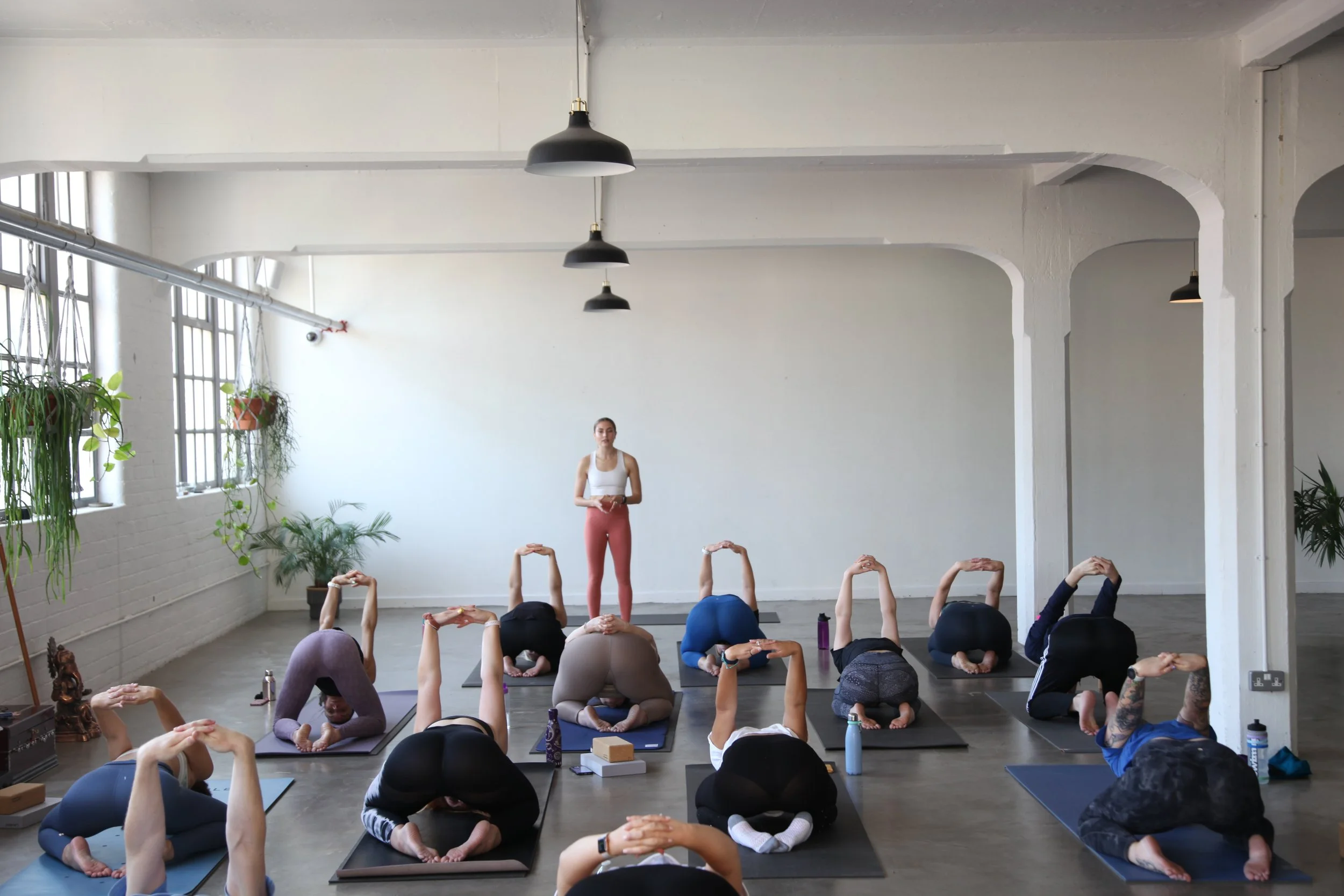 A group of people participating in a yoga class in a bright studio with large windows, plants, and black hanging lights. Evie, our cofounder and instructor standing in front leading the class.