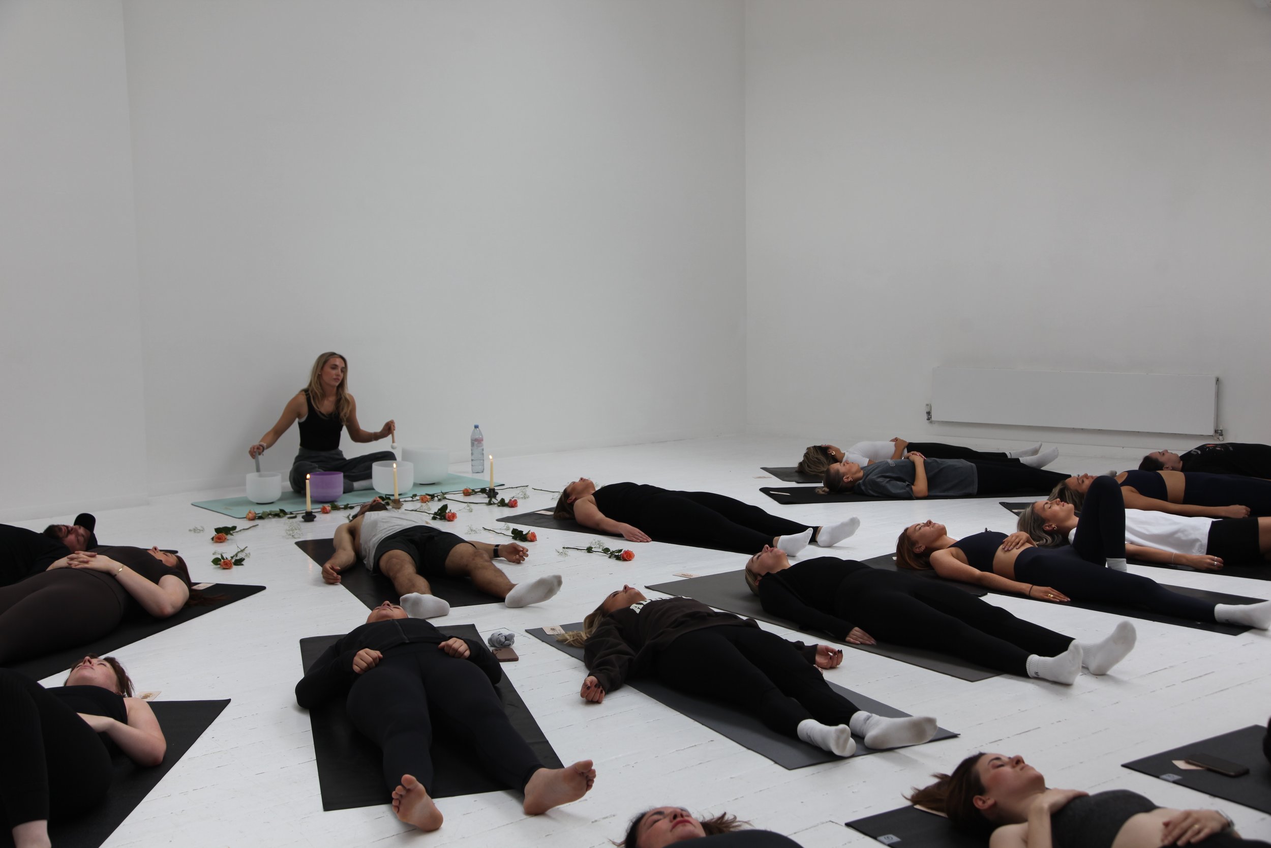 A group of people lying down on yoga mats in a large white room, participating in a soundbath meditation class, with a woman sitting cross-legged at the front near candles, flowers, and musical instruments.