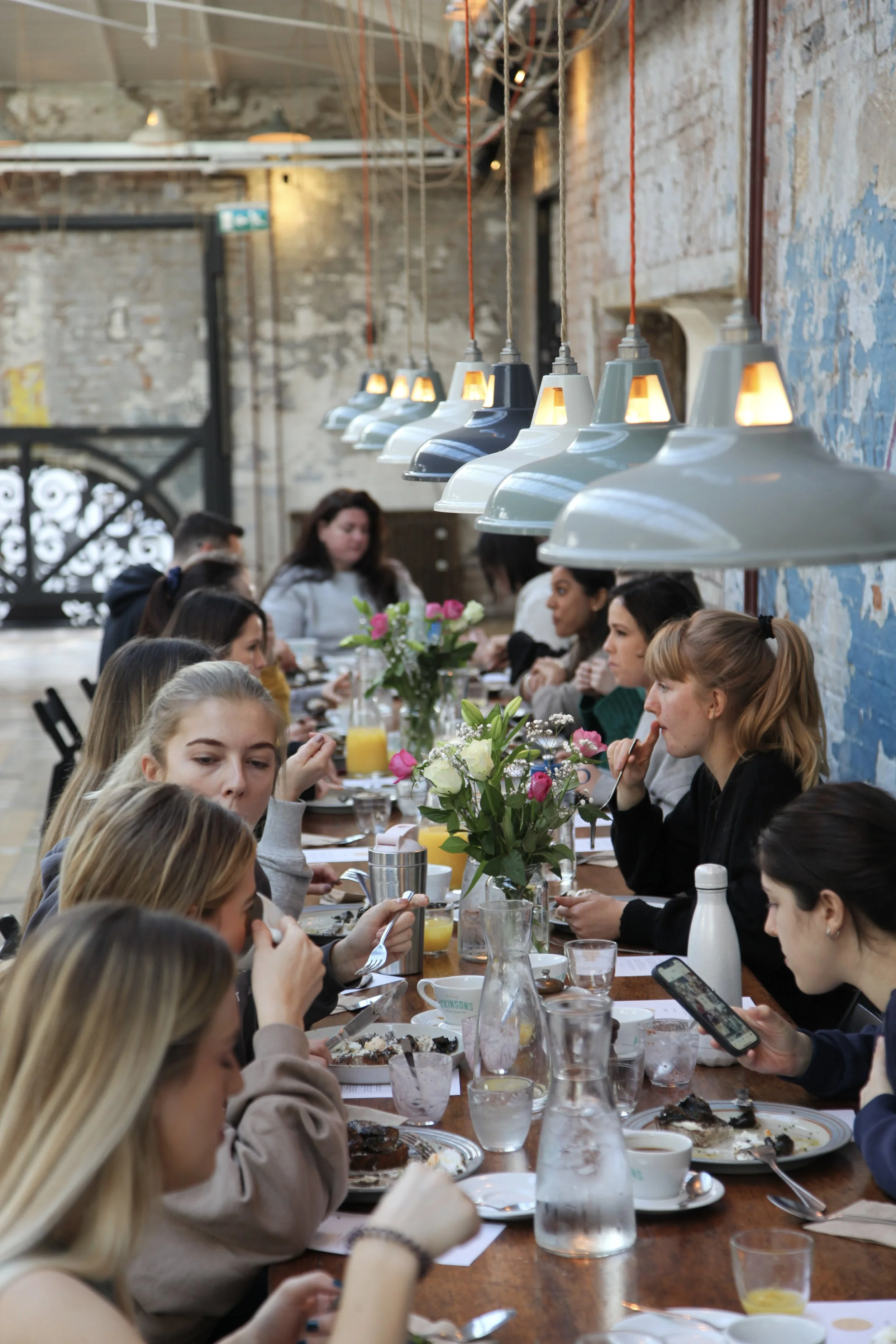 Group of people dining at a long wooden table with floral centerpiece and breakfast dishes inside a rustic, industrial-style restaurant.