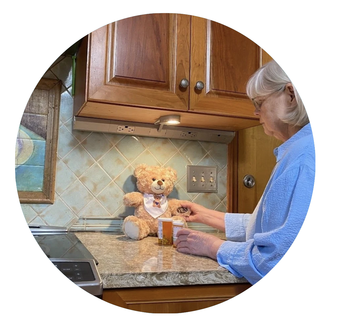 An elderly woman with gray hair and glasses in a blue shirt is in a kitchen, looking at a teddy bear on the counter. The bear is wearing a white bib with a small bear design. There are medicine bottles on the counter and a teddy bear display in the background.