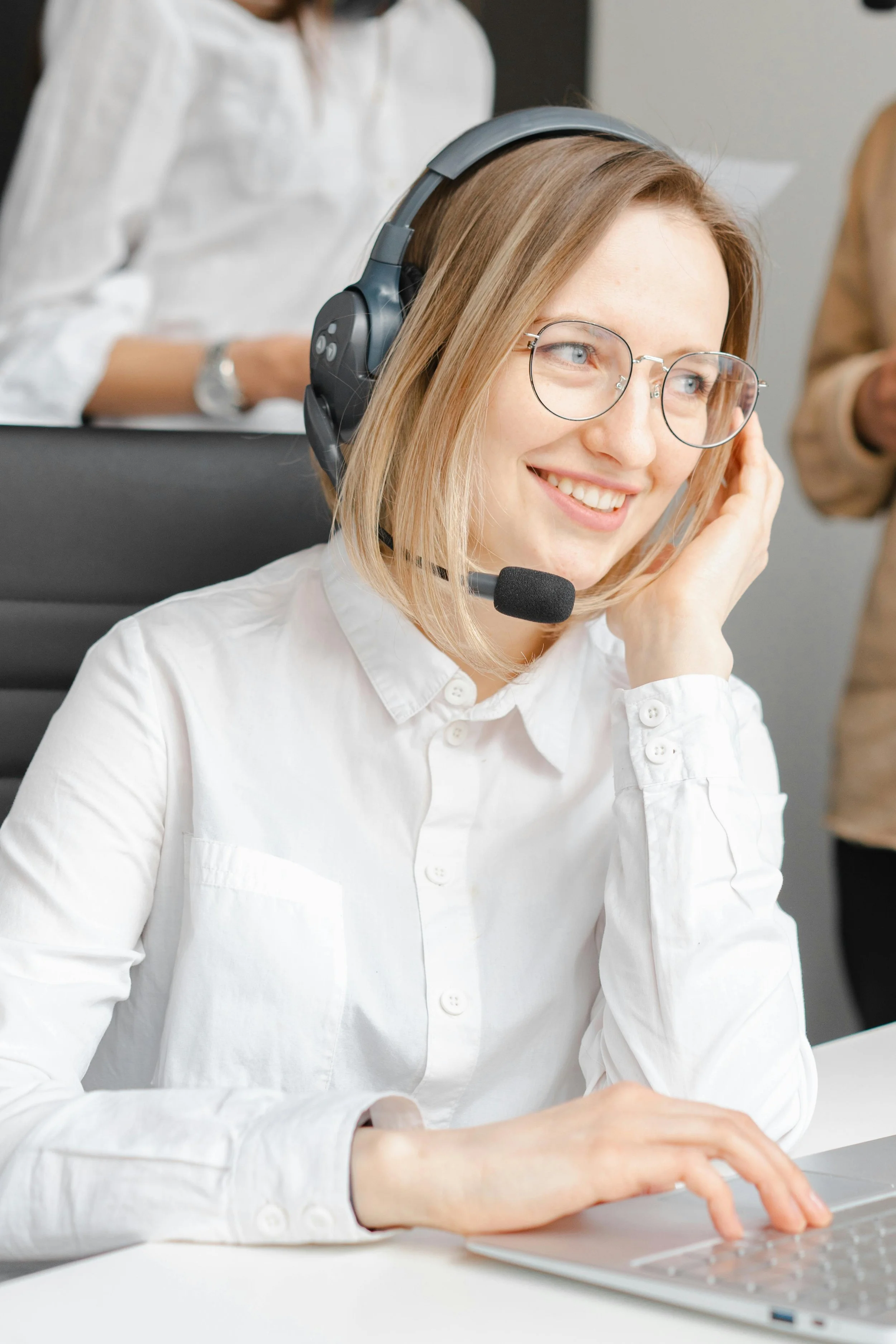 A young woman with blonde hair, wearing glasses, a headset, and a white shirt, smiling while working on a laptop.