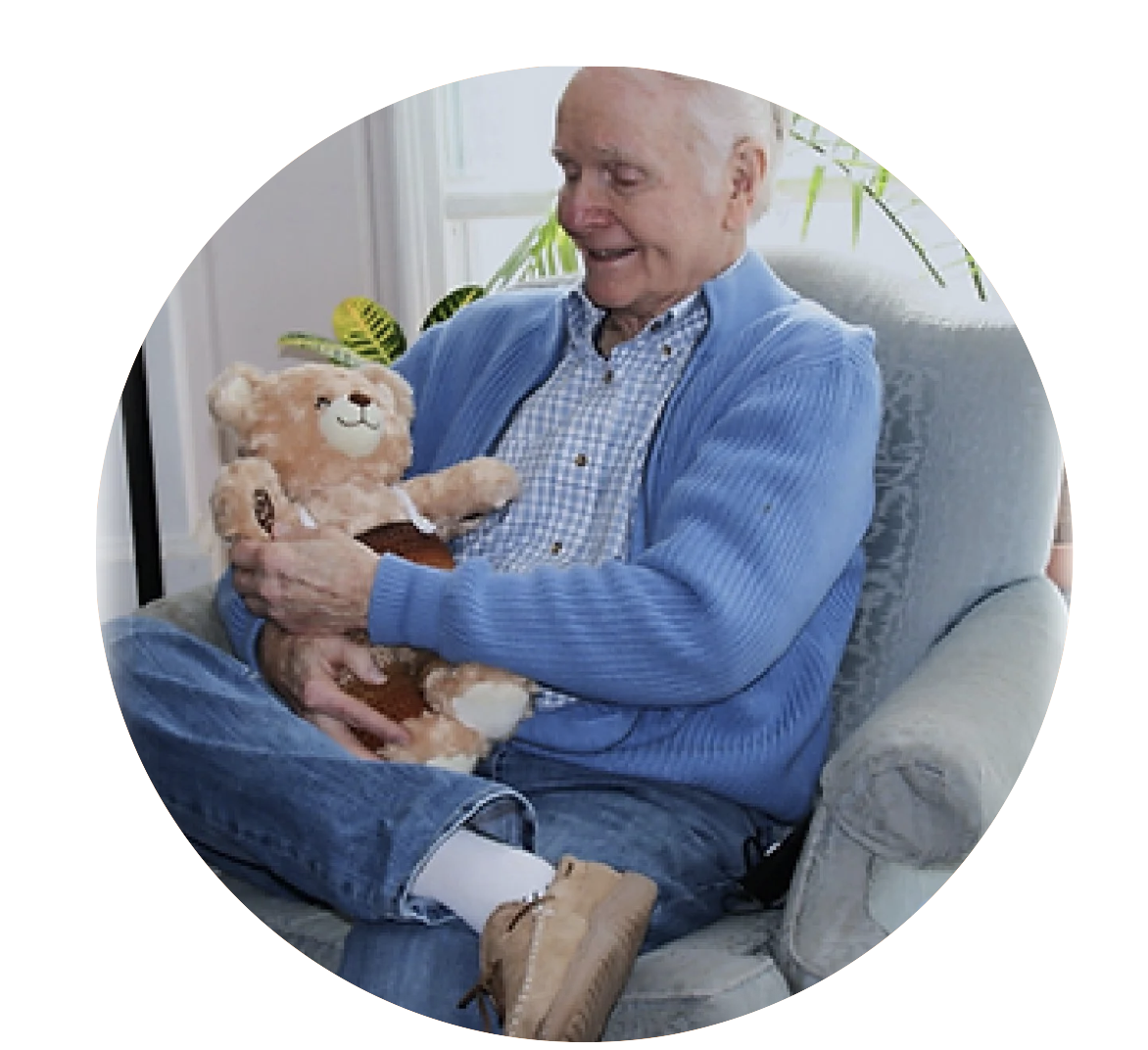 An elderly man sitting on a gray armchair, smiling and holding a teddy bear, in a living room with a window and a plant in the background.
