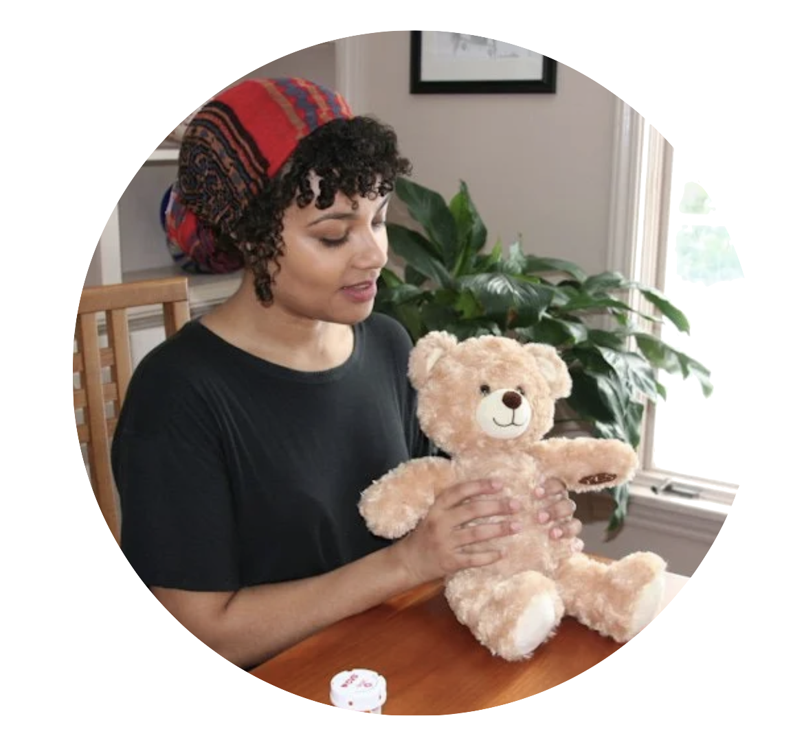 A woman with curly hair wearing a black shirt and a colorful headscarf, sitting at a table, holding and looking at a plush teddy bear.
