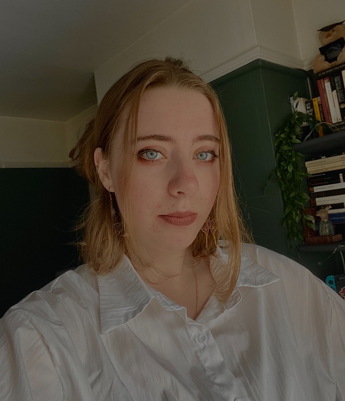 A young woman with light brown hair and blue eyes taking a selfie indoors, wearing a white button-up shirt and gold earrings, with a bookshelf and plant in the background.