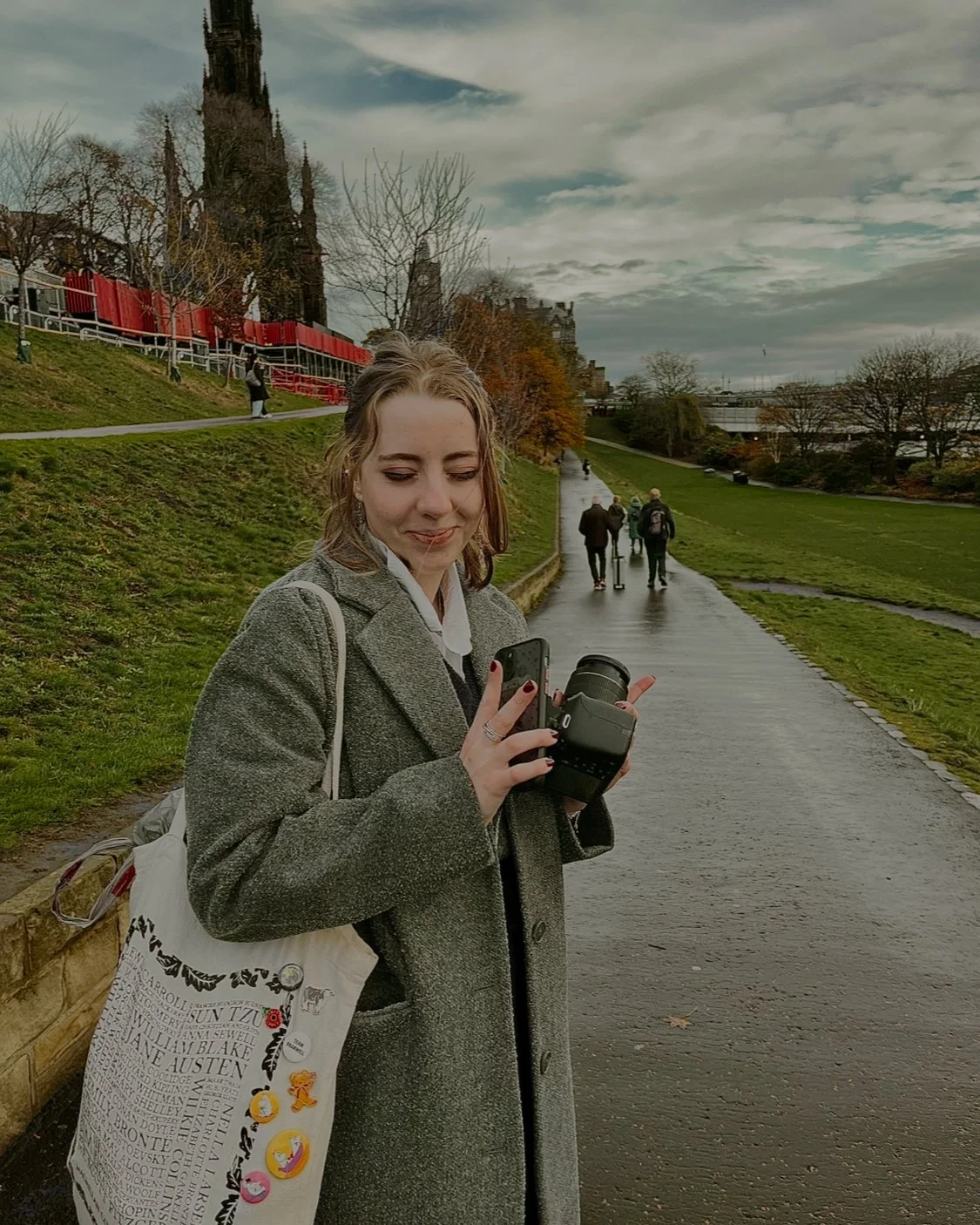 Young woman in gray coat holding a camera and a phone standing on a park pathway, with trees, grass, and other people walking in the background.