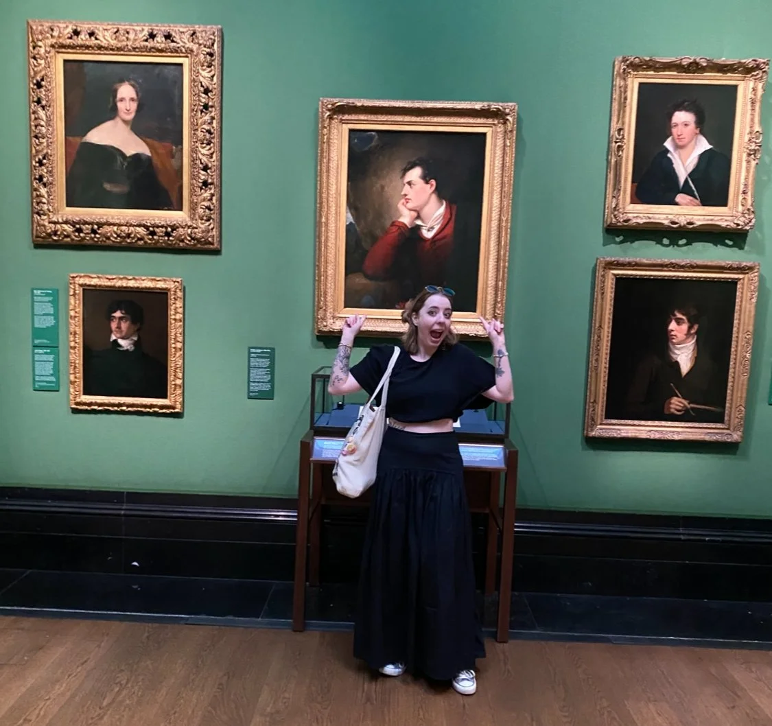 A young woman with tattoos and a white bag standing in front of five framed portraits in an art gallery, making a playful expression with her arms raised and pointing at the portraits.