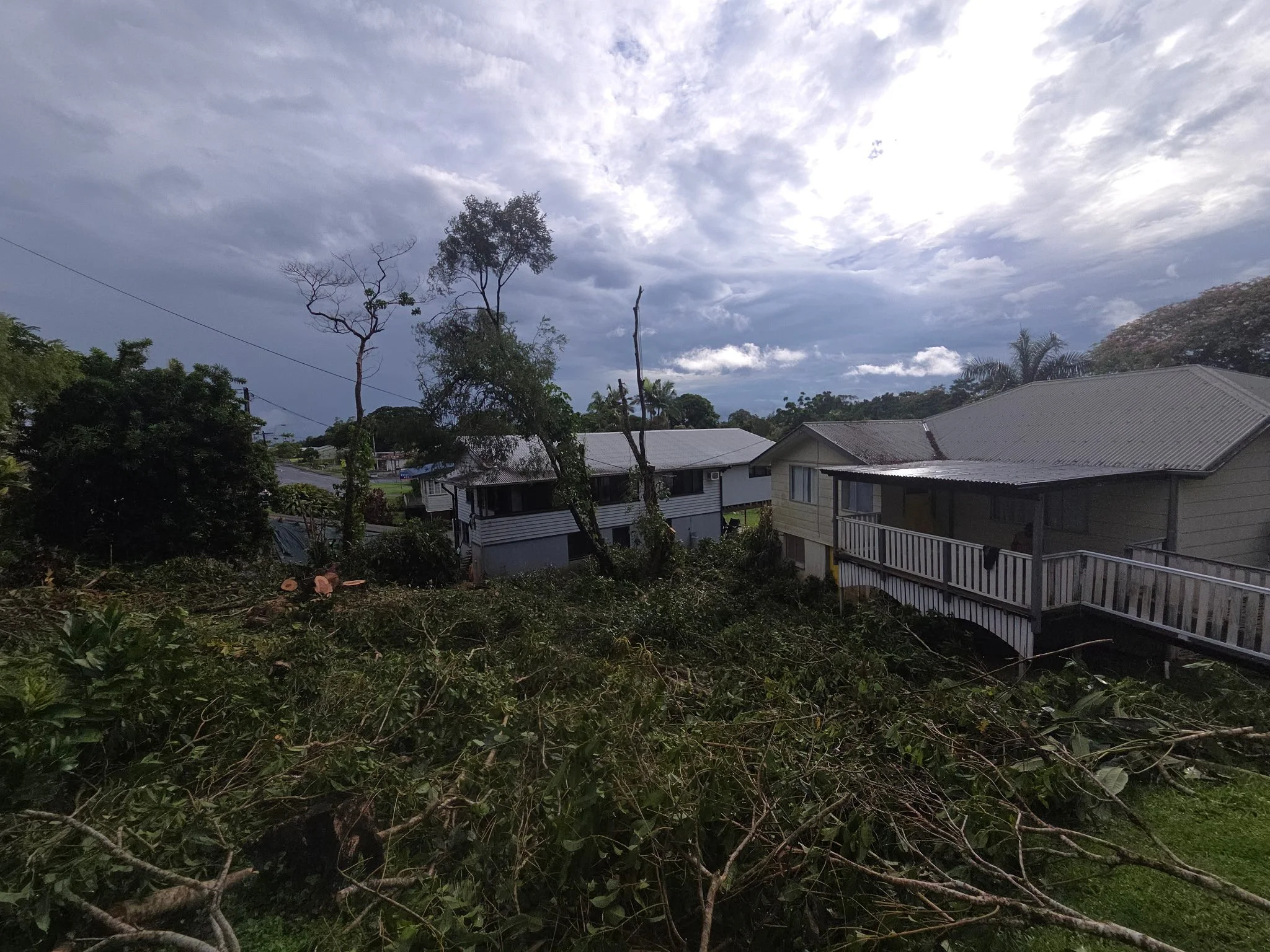 A house with a balcony is surrounded by fallen trees and branches after a storm, with dark clouds in the sky.