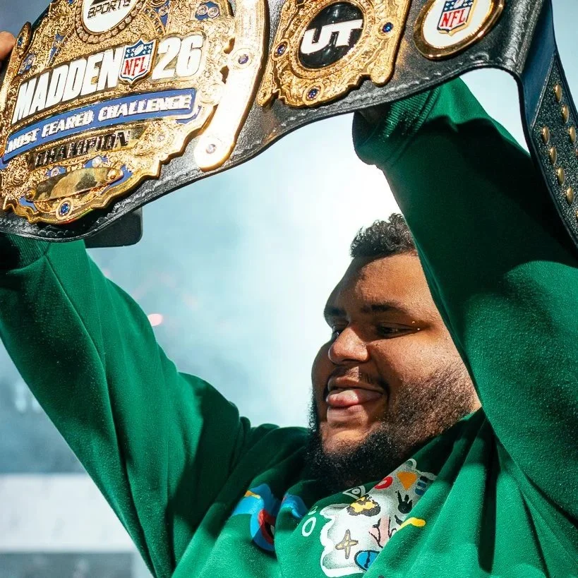 Jonbeast Madden Professional sticking out his tongue while holding up a large NFL championship belt above his head. The belt has the Madden NFL 26 logo and intricate gold designs.