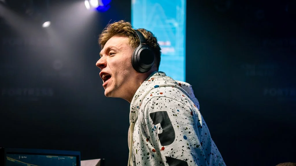 A man with short brown hair wearing large headphones and a white hoodie with colorful paint splatters, speaking passionately at a gaming or e-sports event.
