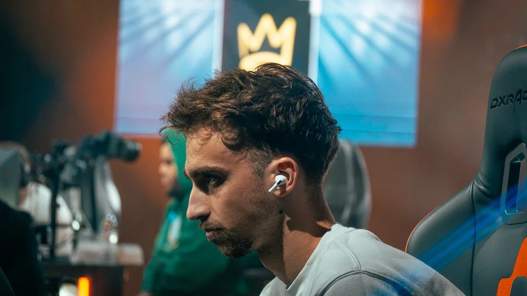 A young man with short, curly brown hair and a light beard, wearing white earbuds, sitting in a gaming or esports arena with a gaming chair. In the background, there is a screen displaying a yellow crown logo on a blue background.