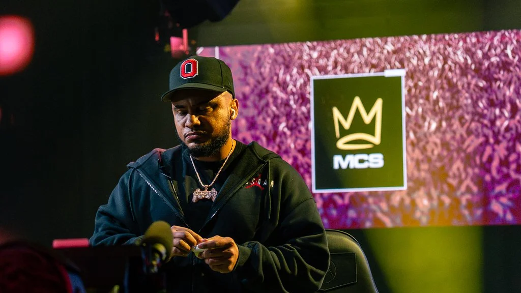 A man wearing a black hoodie and a black Ohio State baseball cap is sitting at a poker table, holding poker chips, in front of a screen displaying a crown logo and the letters 'MCS'.