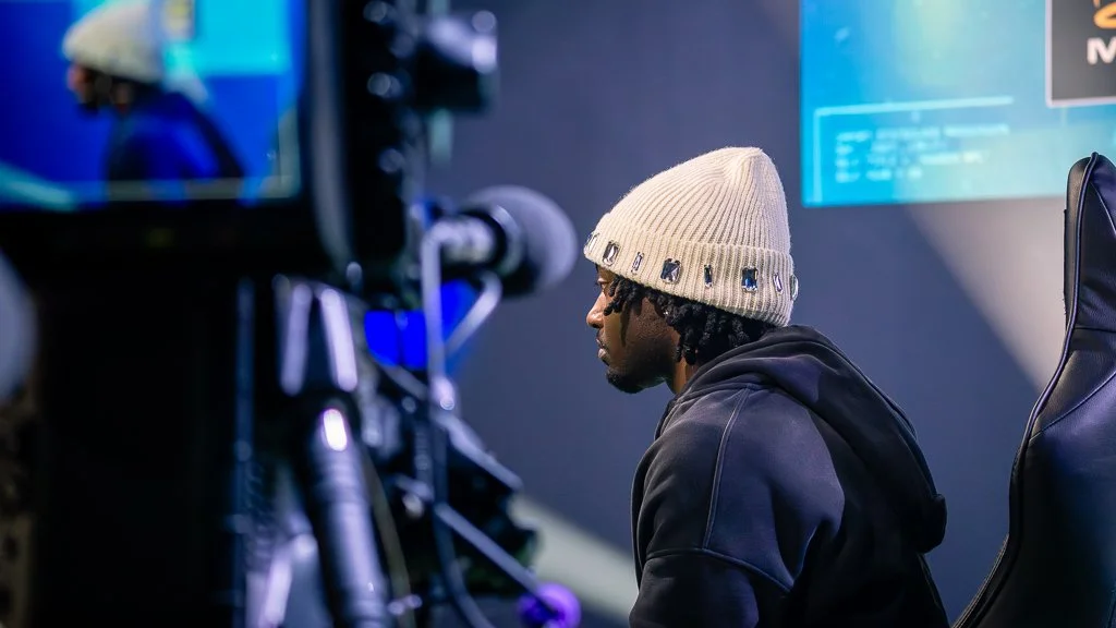 A young man in a beige knit beanie and black hoodie sitting in front of a camera in a studio or recording setting.