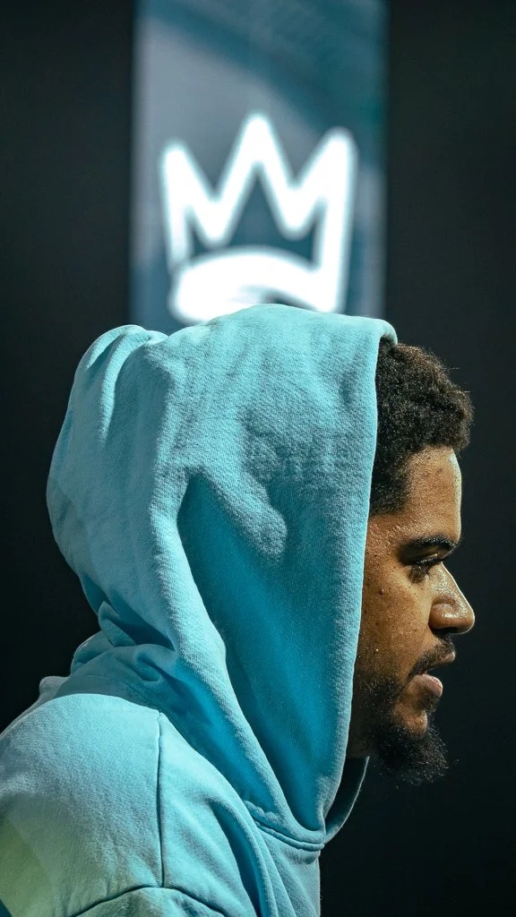 A man wearing a light blue hoodie with the hood up, looking to the right, with a blurred background showing a crown symbol.