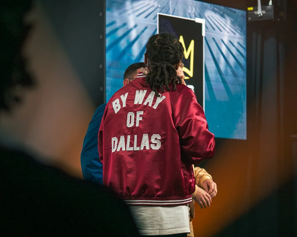 A person with dreadlocks wearing a red jacket with white lettering that says "By Way of Dallas" is standing in front of a large screen or display at an event or conference.