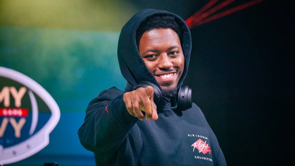 Young man wearing a hoodie and headphones, smiling and pointing at the camera.
